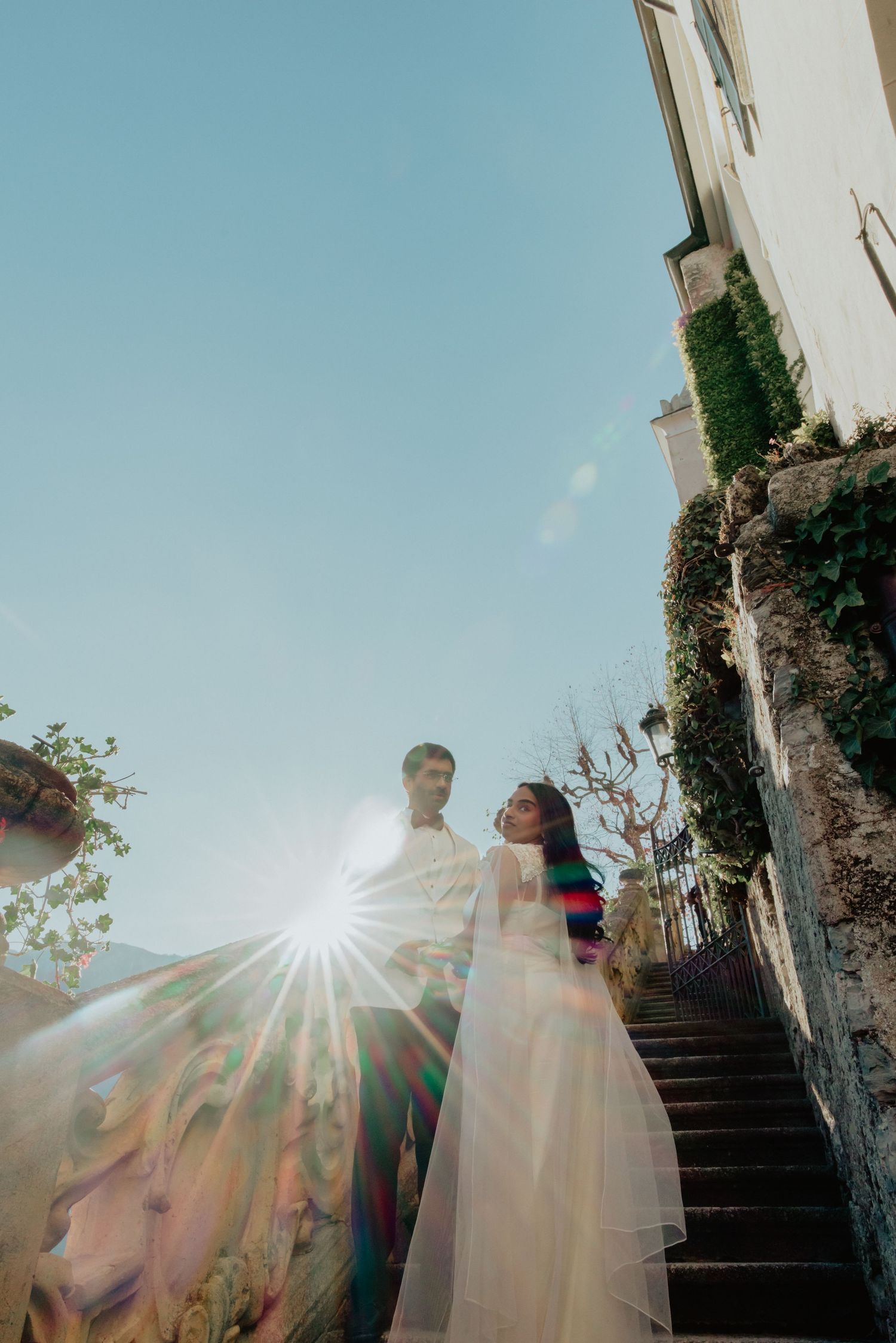 Wedding couple standing on sunlit stone stairs with lens flare, ivy-covered walls, and blue sky background.