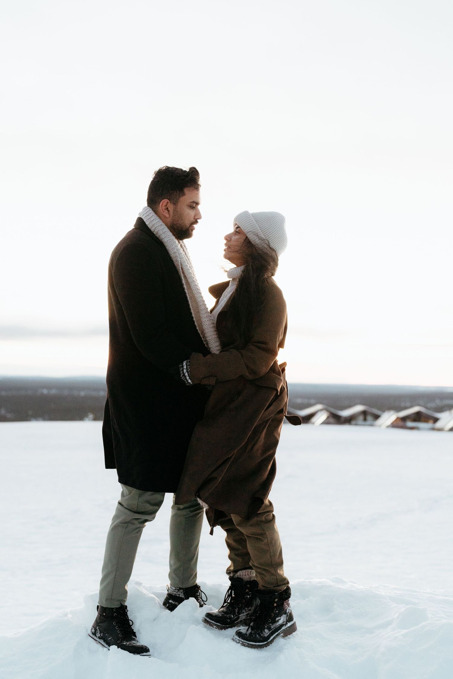 Couple embracing in snowy winter landscape, wearing brown coats and boots, sharing a romantic moment outdoors.