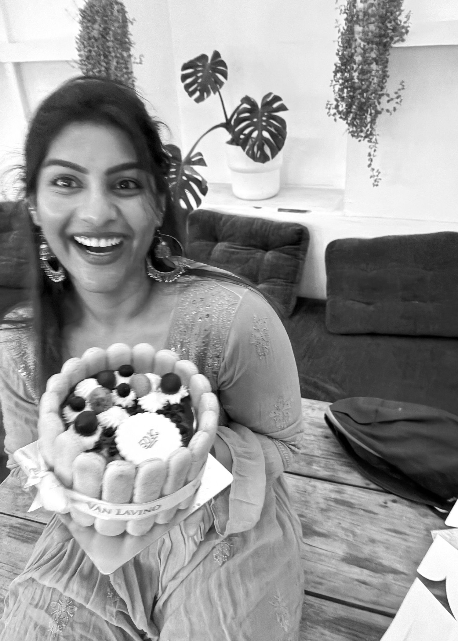 Smiling woman holding a decorated cake in a black and white photo with monstera plants in the background.
