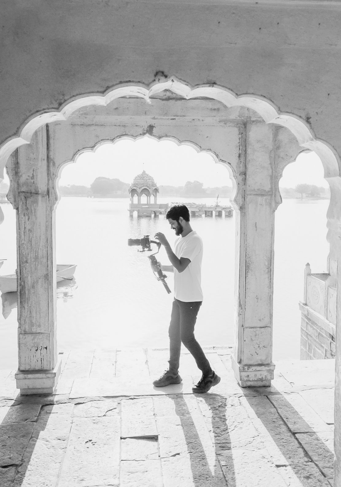 Black and white photo of a man filming with a gimbal stabilizer under an ornate arch at Gadisar Lake, Jaisalmer.