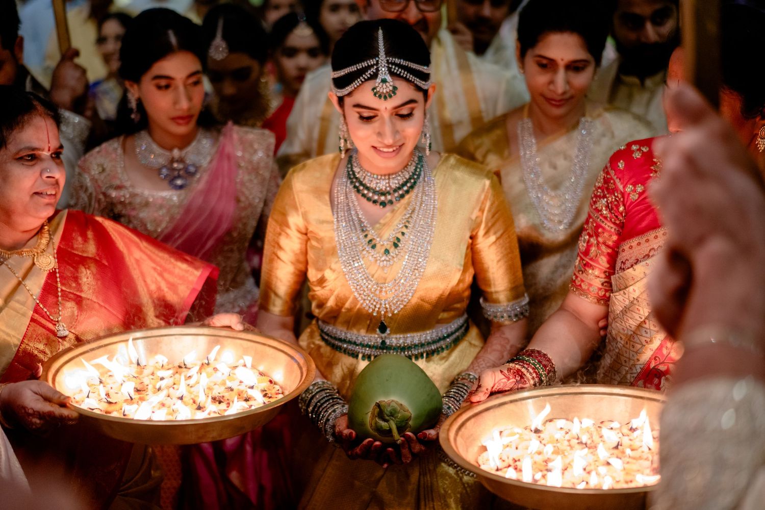 Bride in gold saree holds floral bowl during traditional South Indian wedding ceremony with guests in colorful attire.