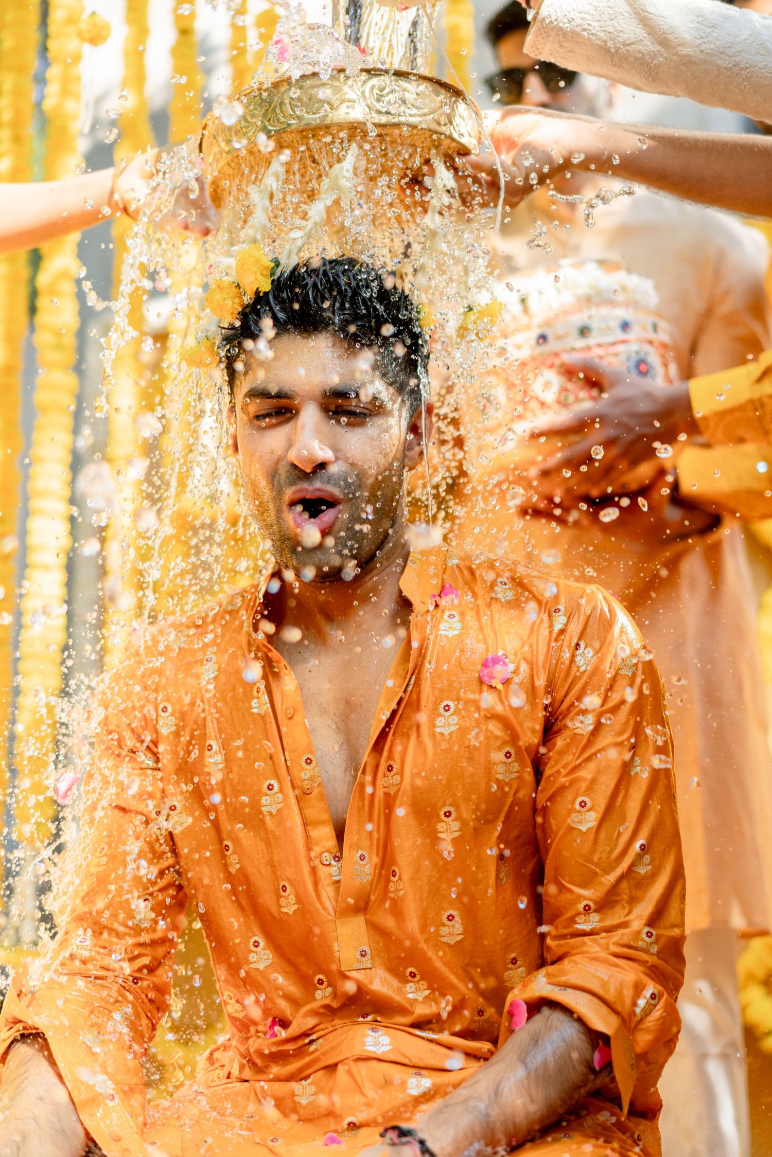 Man in orange kurta has water poured over his head during a traditional Indian Haldi ceremony.