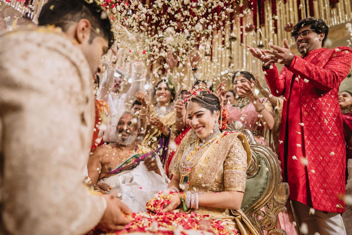A joyful Indian bride in gold bridal wear during a vibrant wedding ceremony with flower petals raining down.
