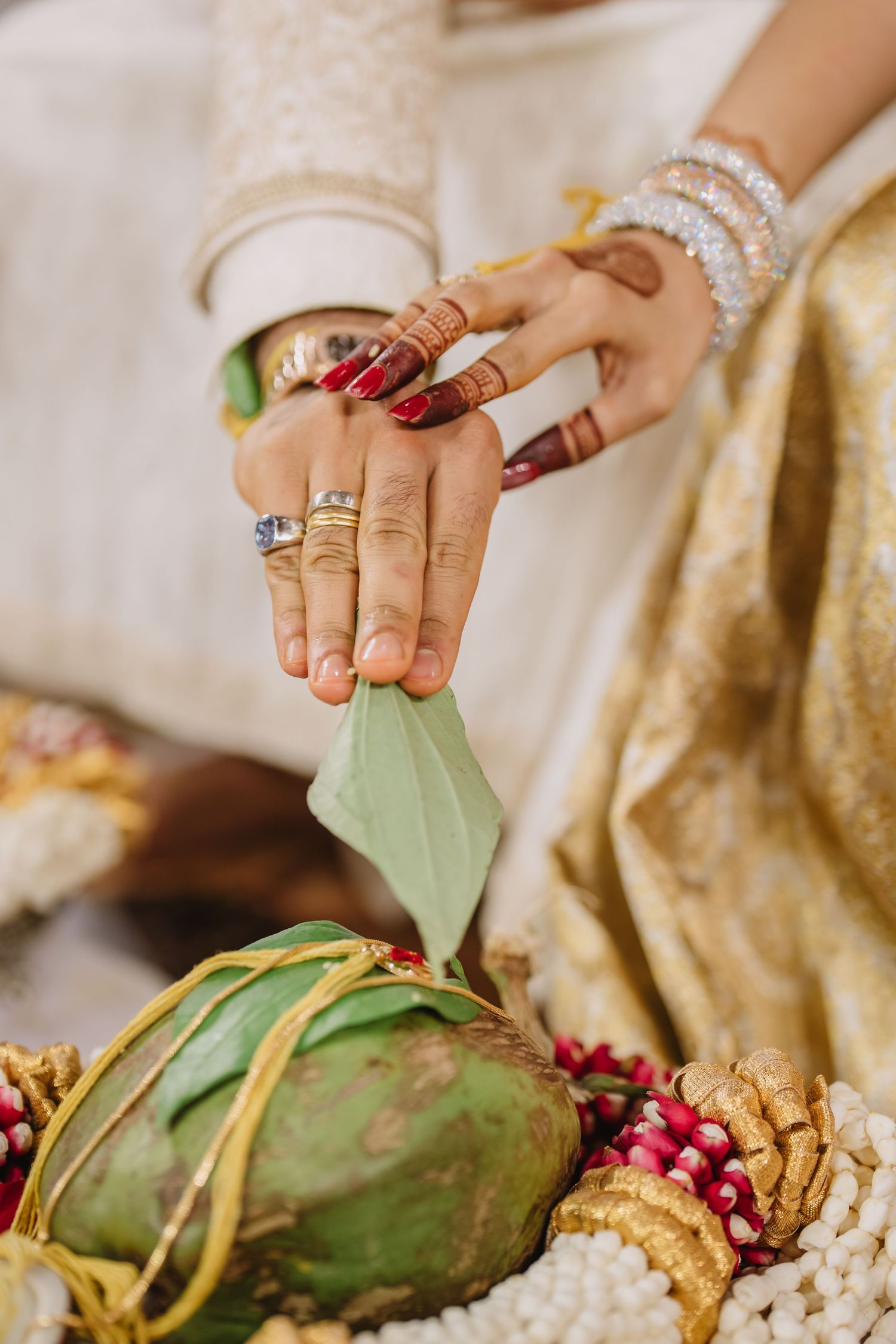 Close-up of bride and groom hands during Indian wedding ceremony with henna, bangles, and rings holding a green leaf.