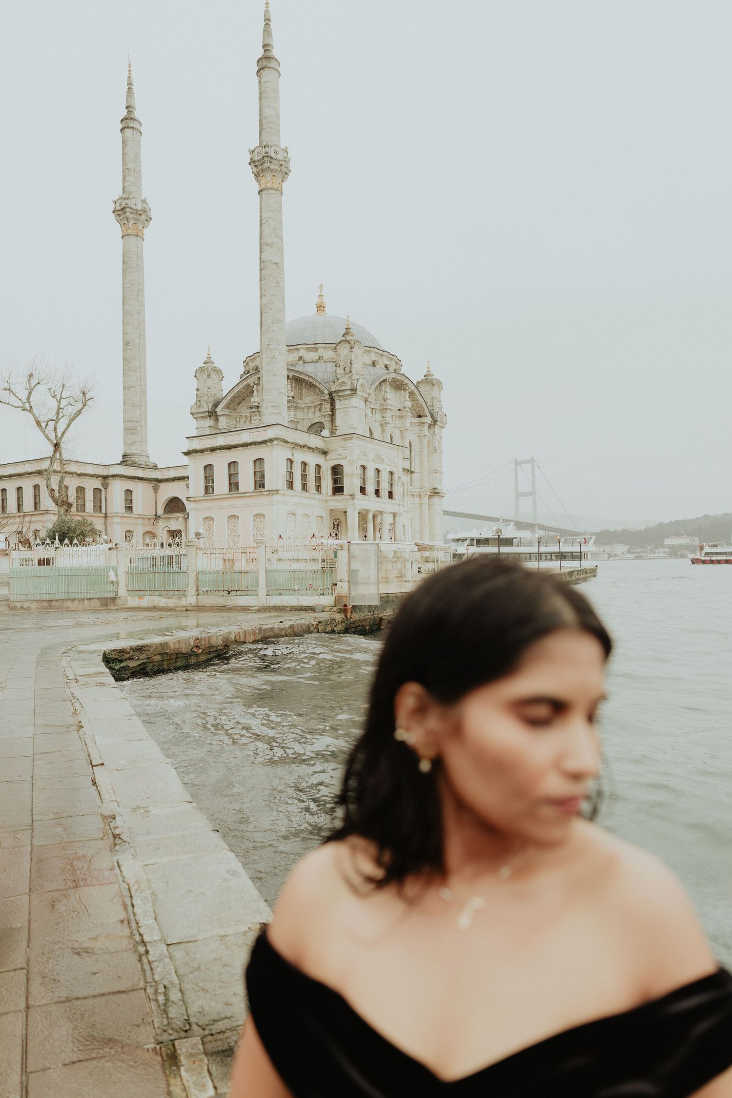 Woman in black off-shoulder dress with Ortakoy Mosque and Bosphorus Bridge in Istanbul.
