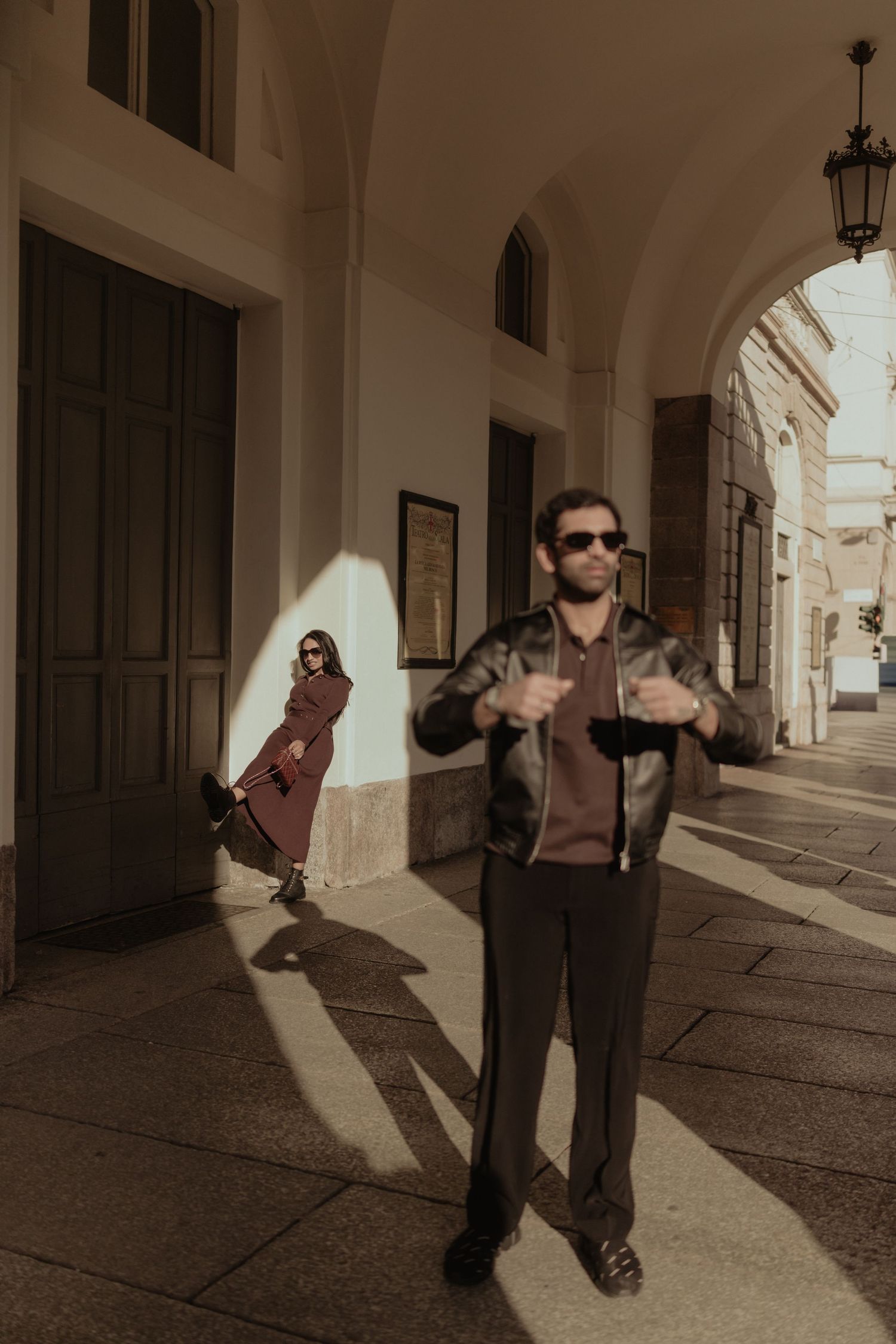 Fashionable couple in urban arcade, man in leather jacket with sunglasses, woman in brown coat in background.