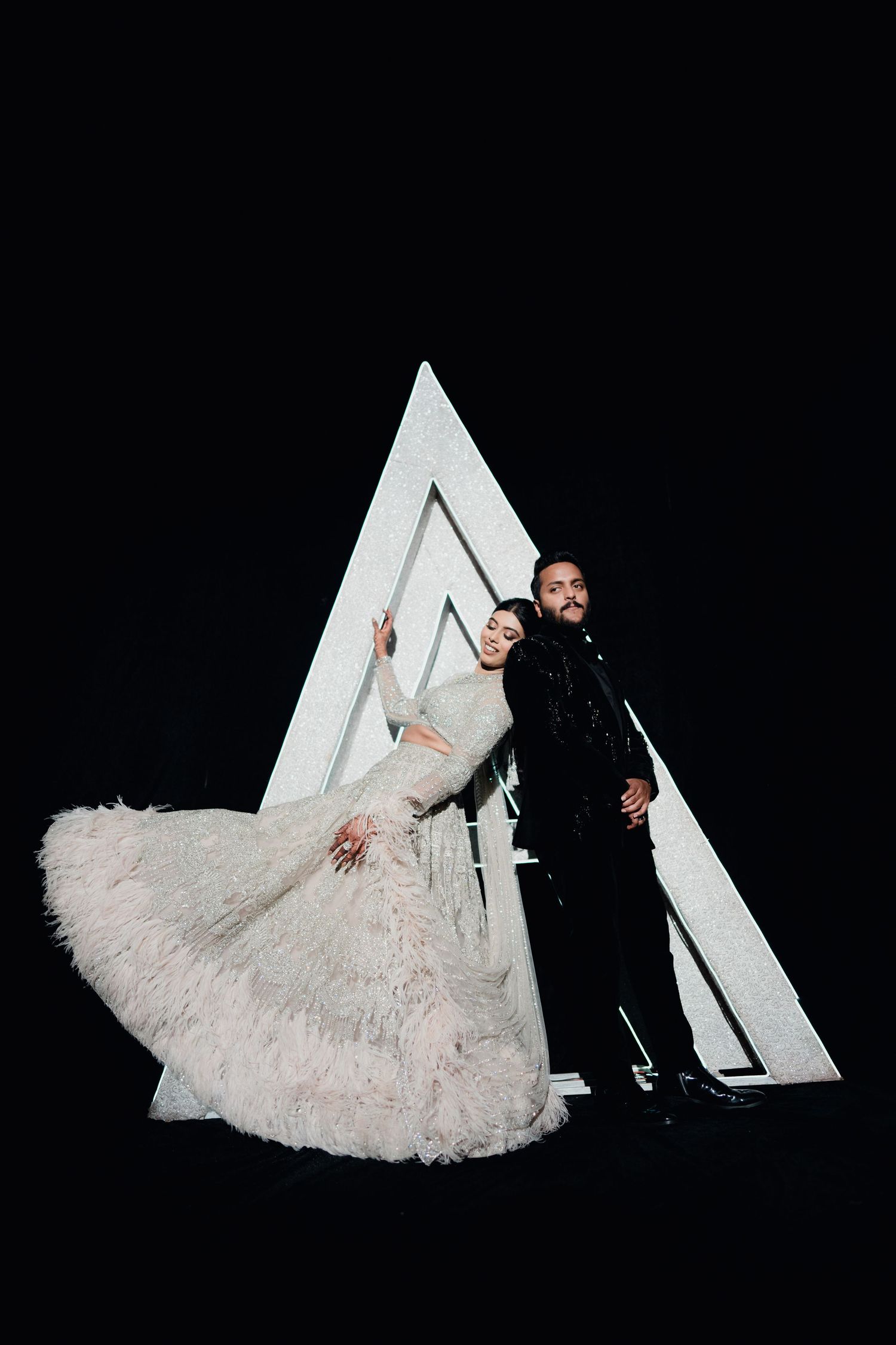 Bride in feathered white gown and groom in black suit pose before illuminated triangle backdrop.