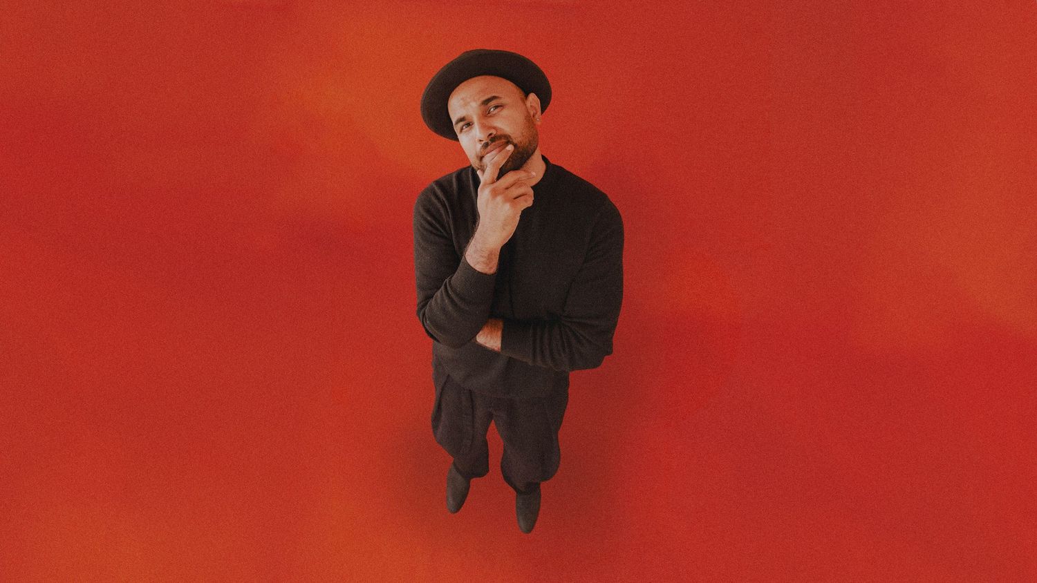 A man in black outfit and hat poses thoughtfully against a vibrant red background, looking upward.