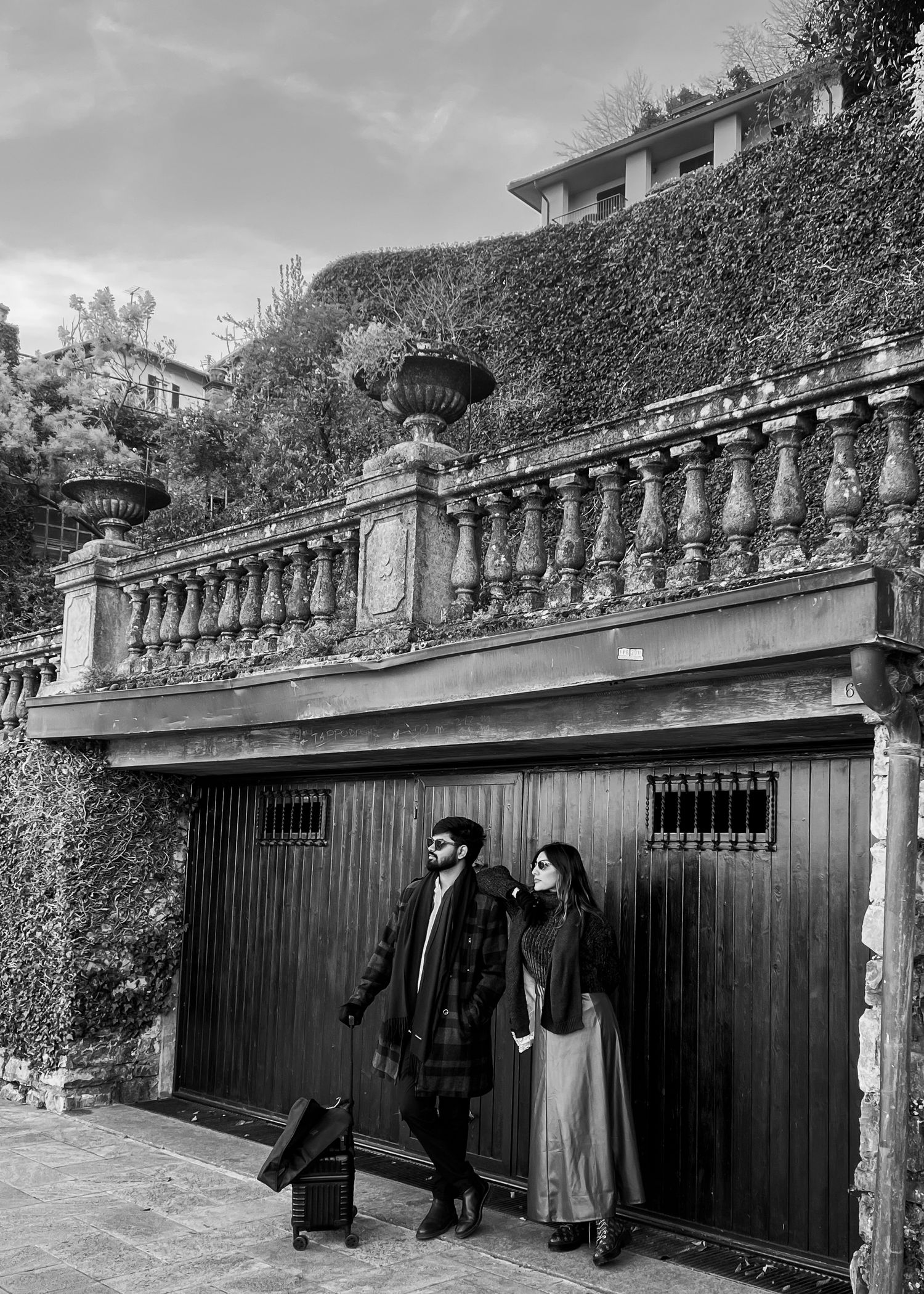 Black and white photo of a couple standing before an ornate stone balustrade with ivy-covered walls and decorative urns.