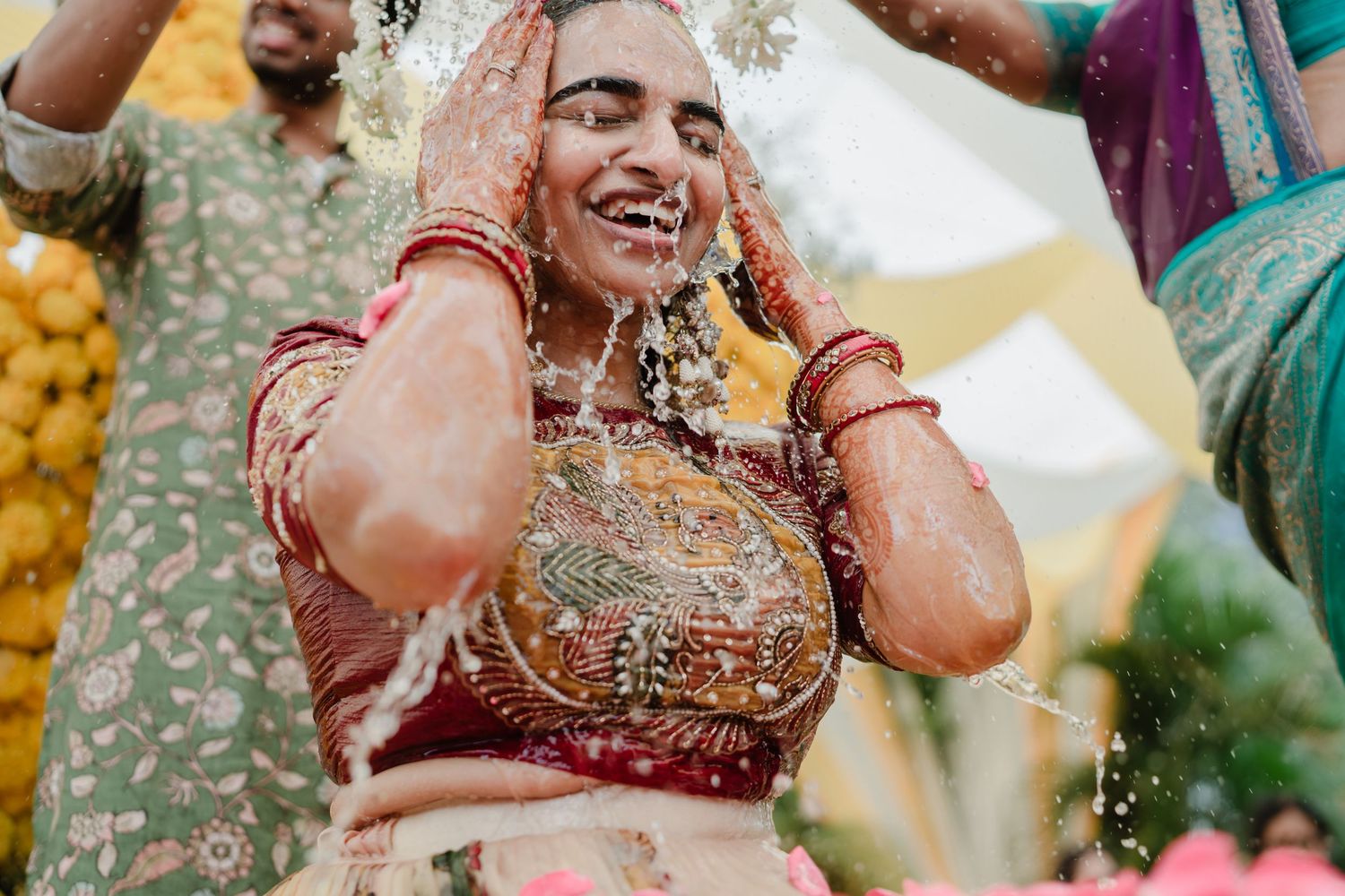 Joyful Indian bride in red lehenga laughing during traditional haldi ceremony, adorned with jewelry and floral decorations.