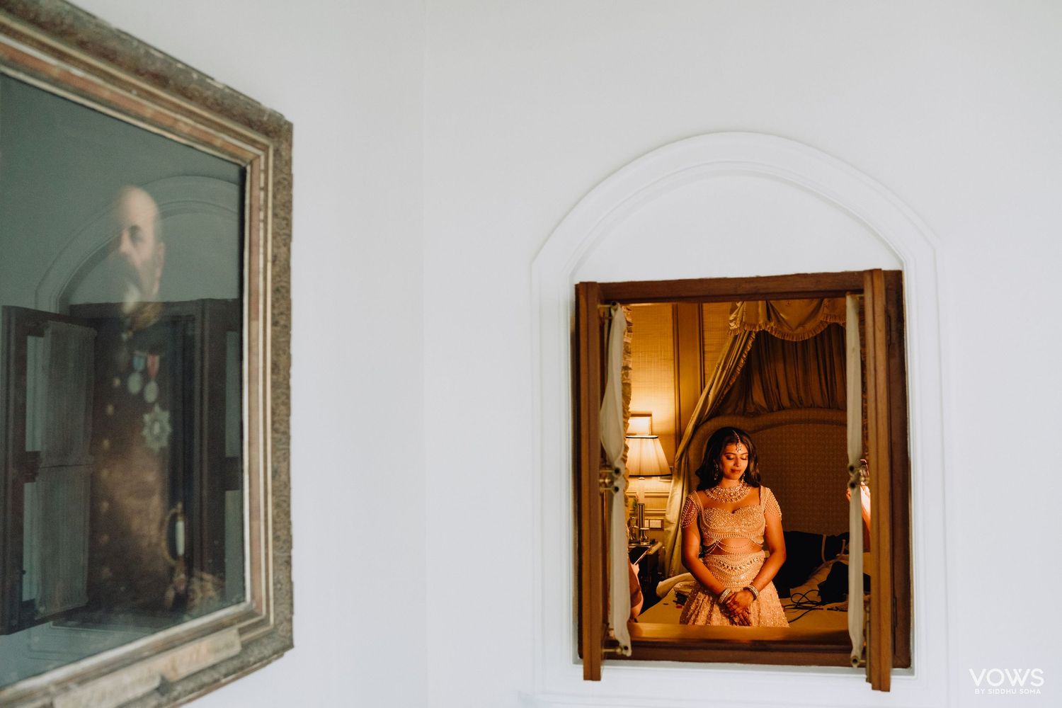 Bride seen in ornate mirror reflection during wedding preparations in an elegantly lit room with vintage portrait on wall.