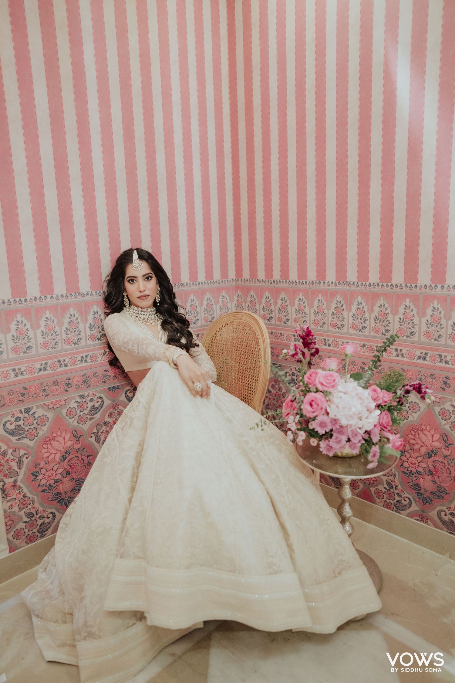 Bride in ivory embroidered lehenga poses gracefully in pink striped room with floral arrangement.