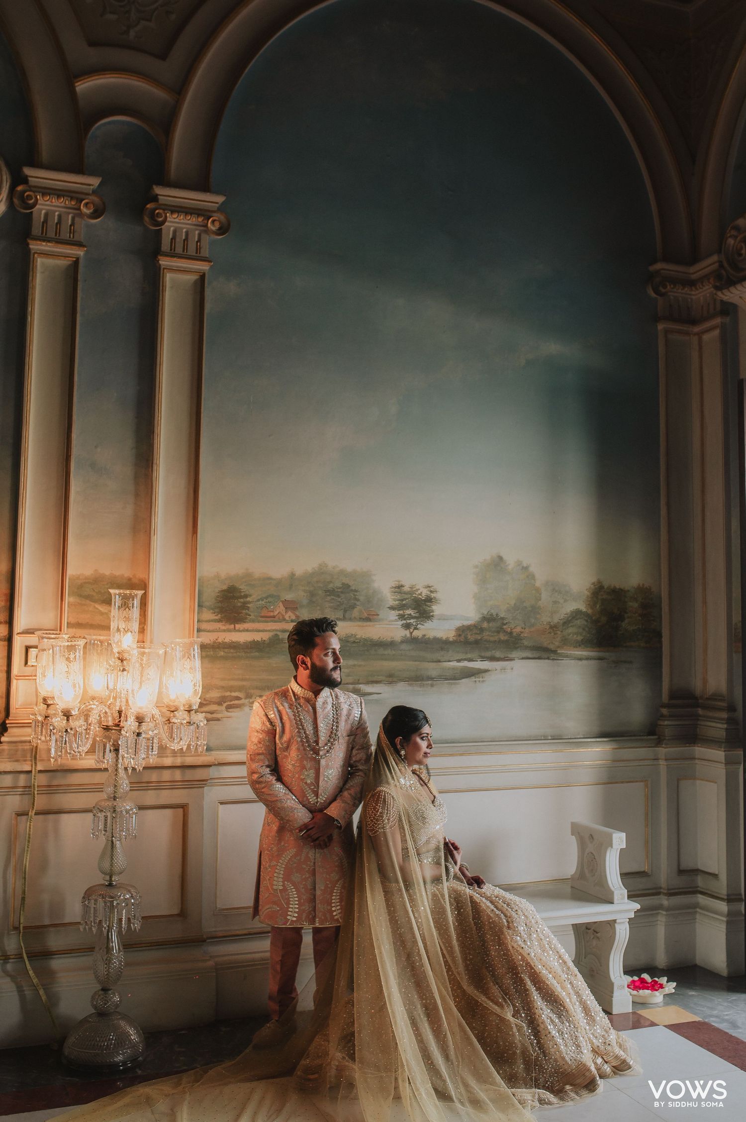 Elegant Indian couple in bridal attire posing in ornate hall with mural backdrop and crystal candelabra.