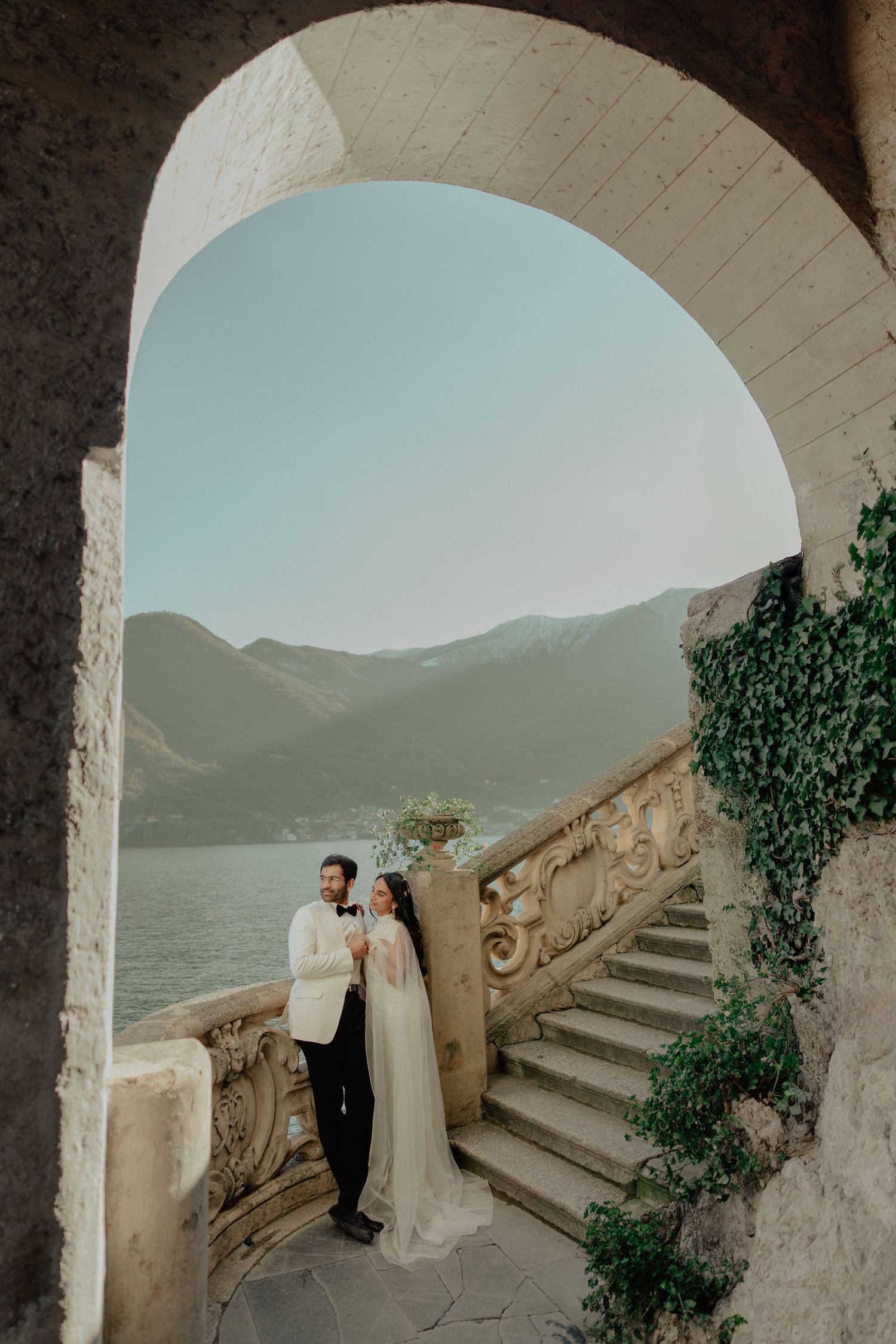 Bride and groom pose under stone arch at Lake Como villa with mountain views and ornate staircase.