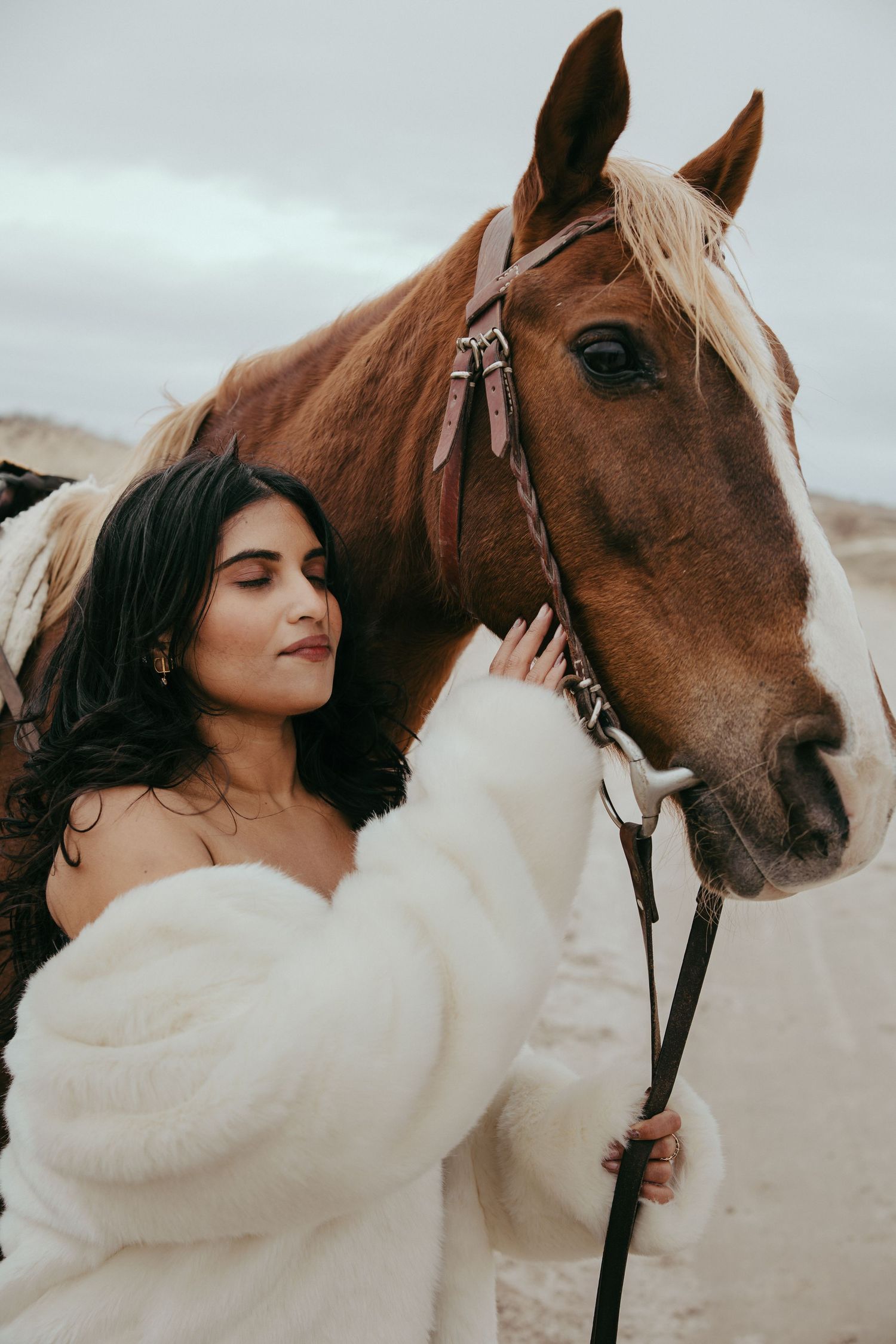 Woman in white fur coat tenderly poses beside a brown horse outdoors.