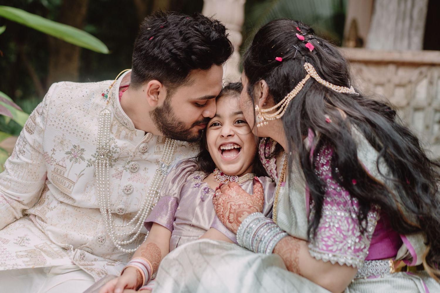 A joyful South Asian family in traditional attire, parents kissing their laughing little girl on the cheeks.