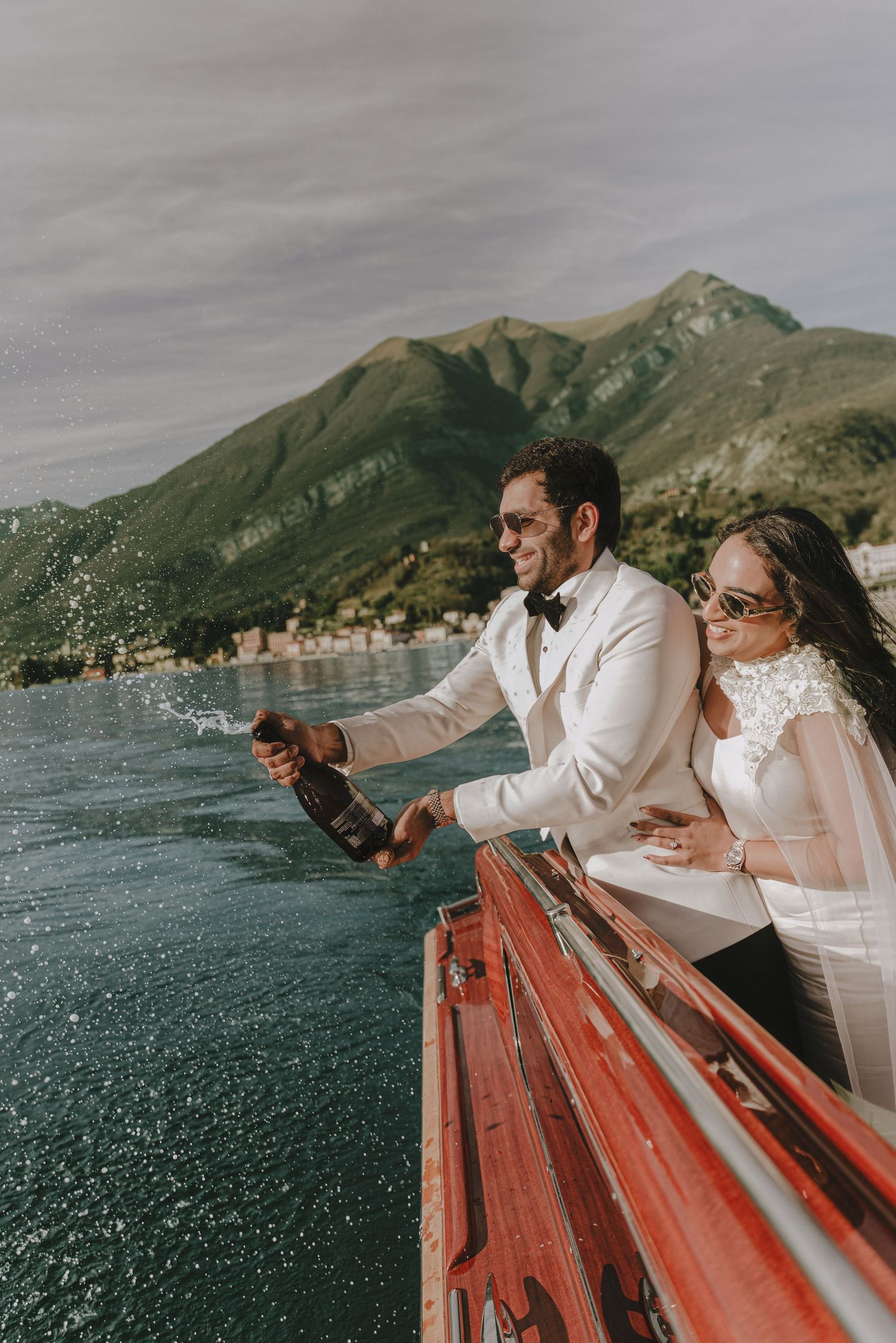 Wedding couple pops champagne off boat bow on Lake Como with mountain backdrop on their wedding day.