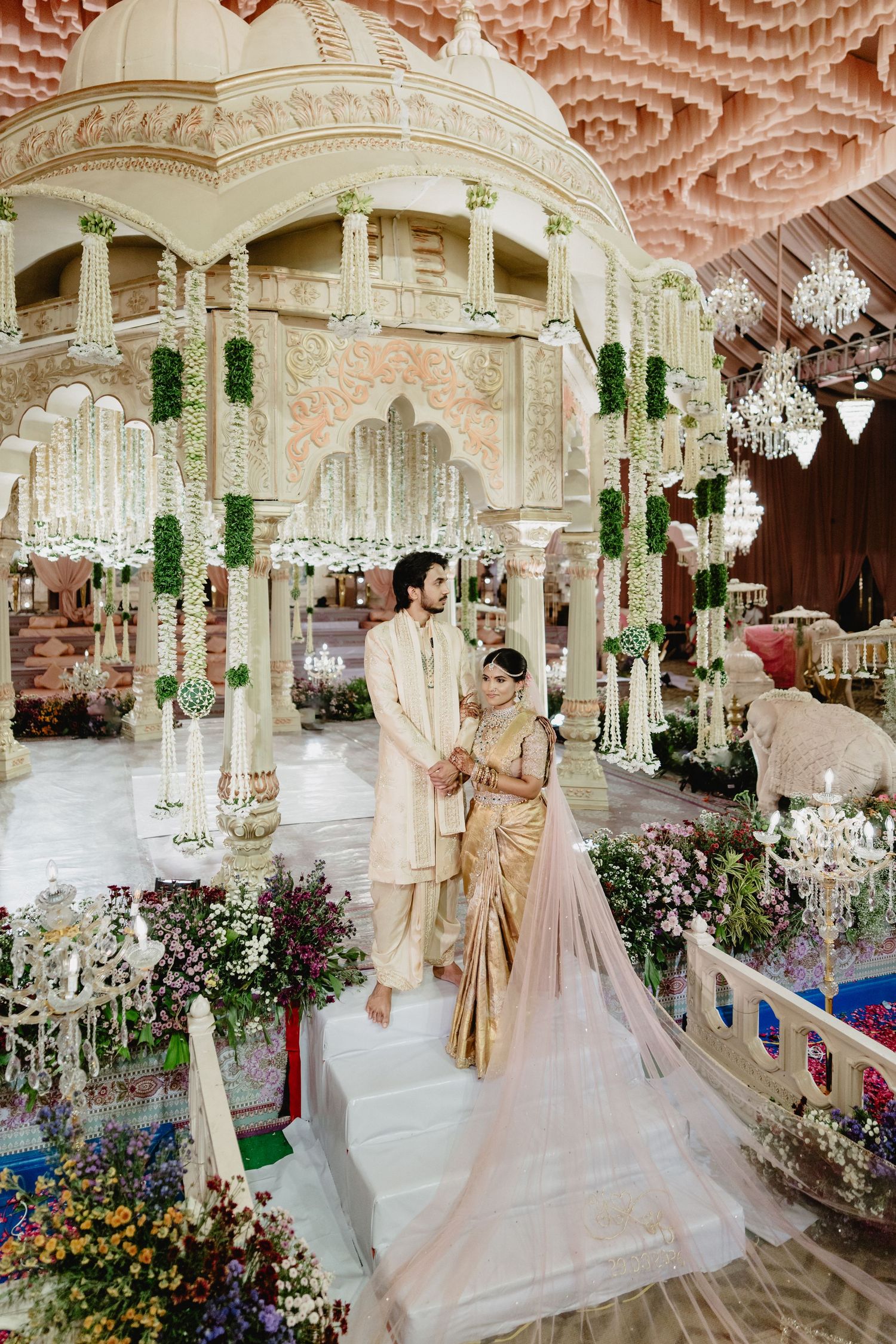 Bride in gold saree and groom in ivory sherwani standing in an ornate mandap with white floral wedding decor.