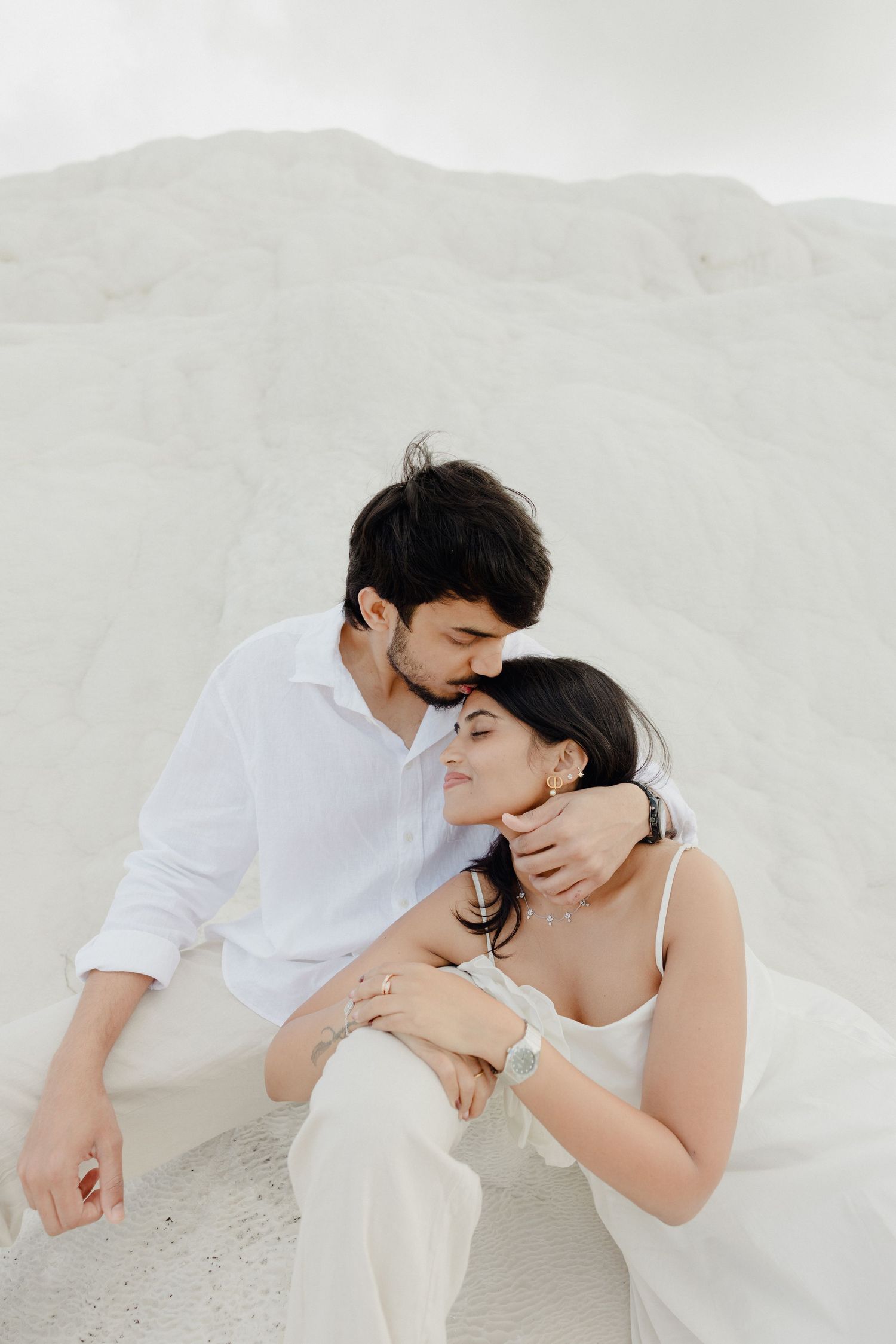 Romantic couple in white outfits sitting together against white sandy dunes.