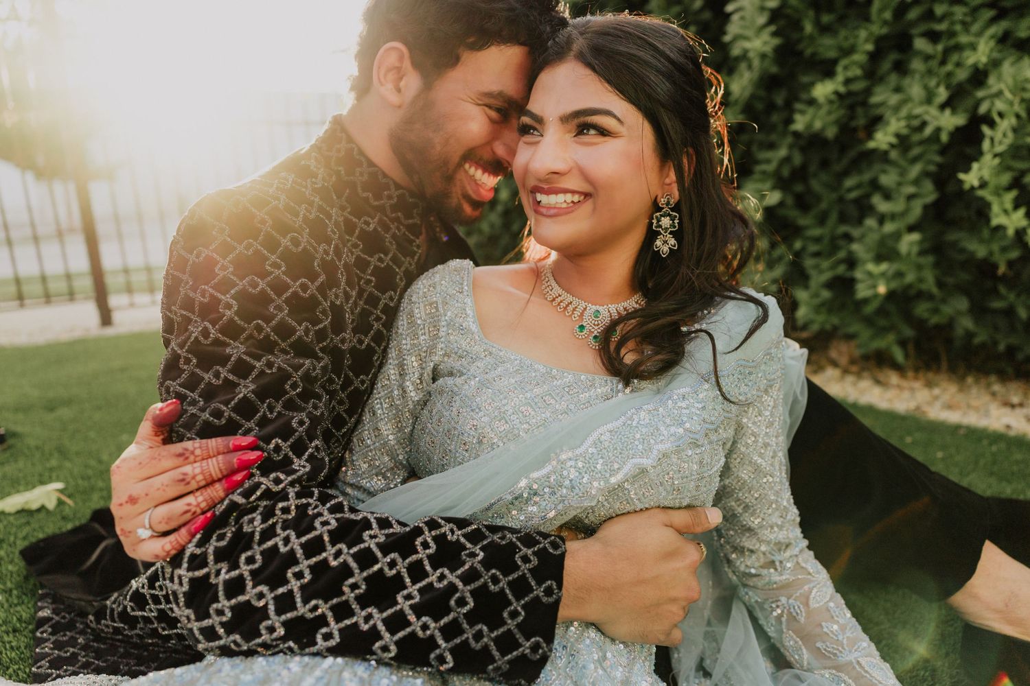 Couple in traditional South Asian attire embracing outdoors, woman in light blue lehenga with gold jewelry, man in black sherwani.