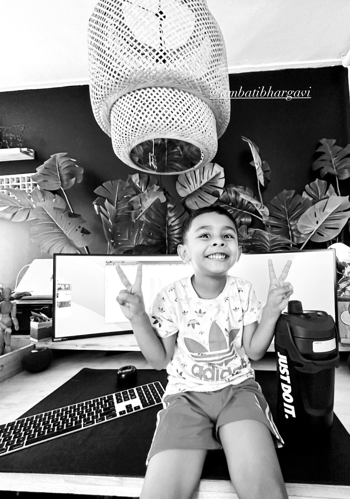 Happy child making peace signs at a desk with monitors, plants, and pendant light in black and white.