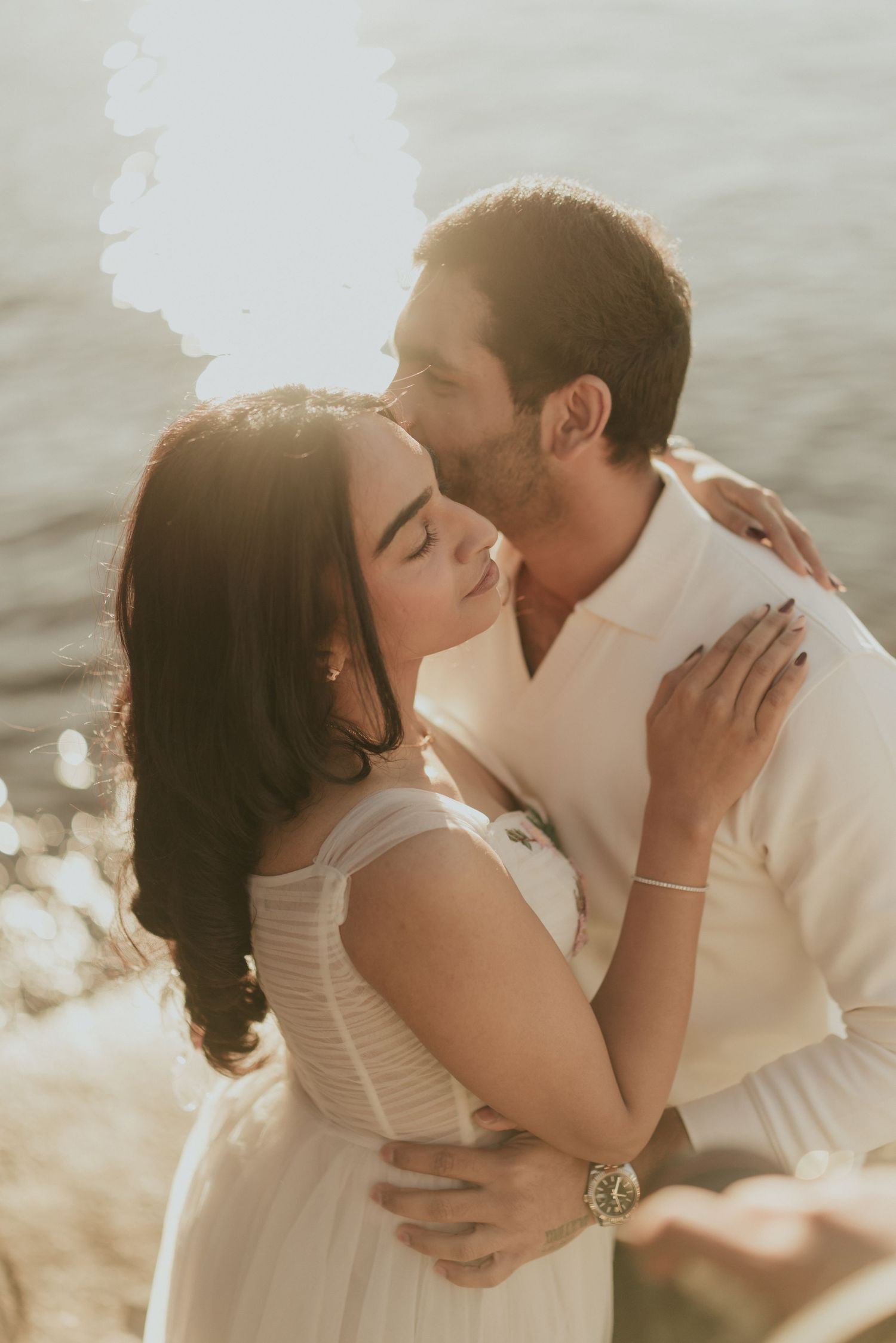 Couple sharing a romantic kiss at golden hour by the water, bride in white dress with bracelet and watch.