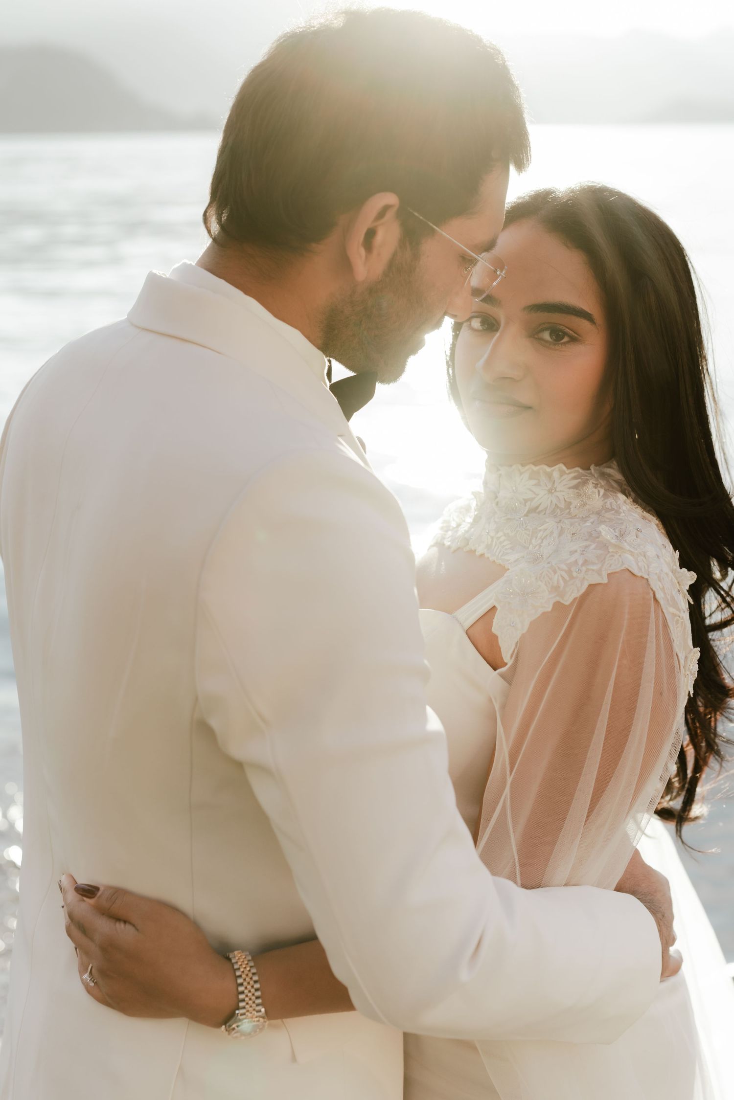 Couple in wedding attire holding each other close with bright sunlight reflecting off the water.