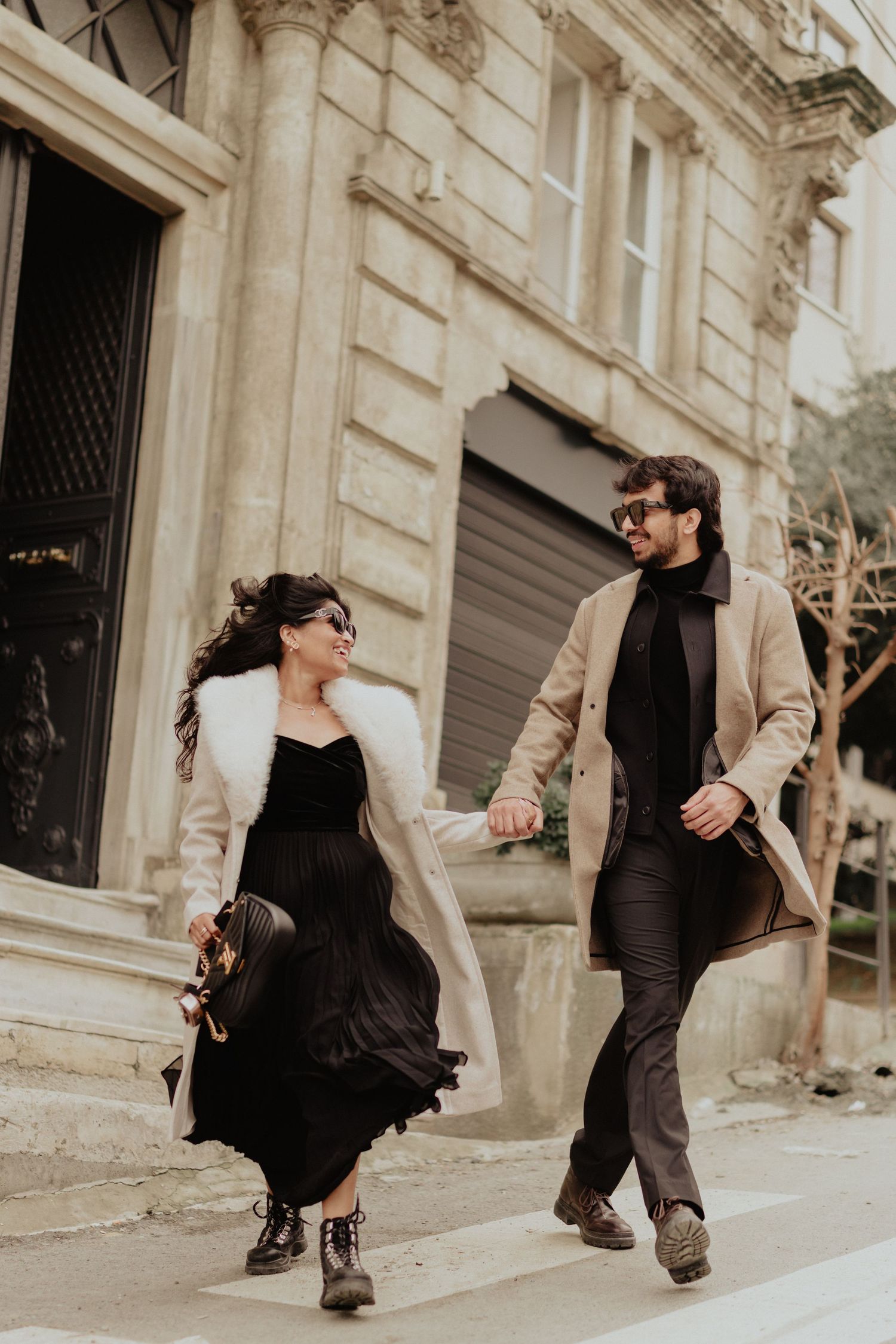 Stylish couple walking hand in hand on a Parisian street, woman in black dress with fur coat, man in beige overcoat.
