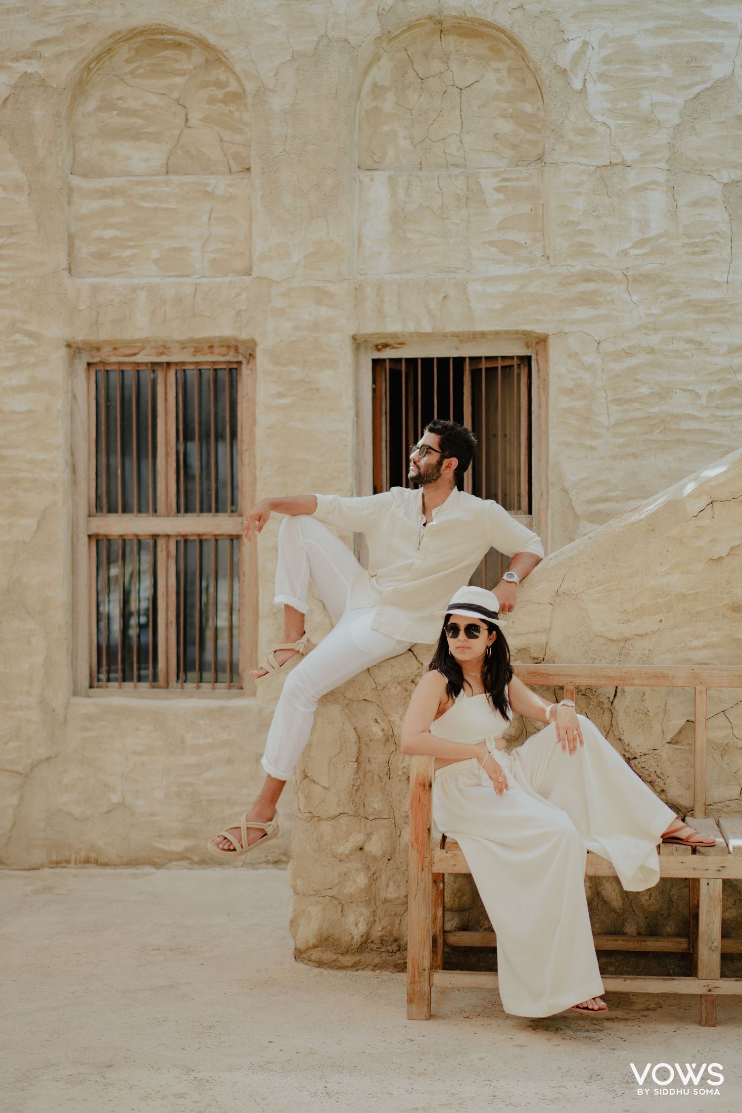 Couple in matching white outfits posing at a rustic stone building, woman seated wearing a hat, man perched on a ledge.