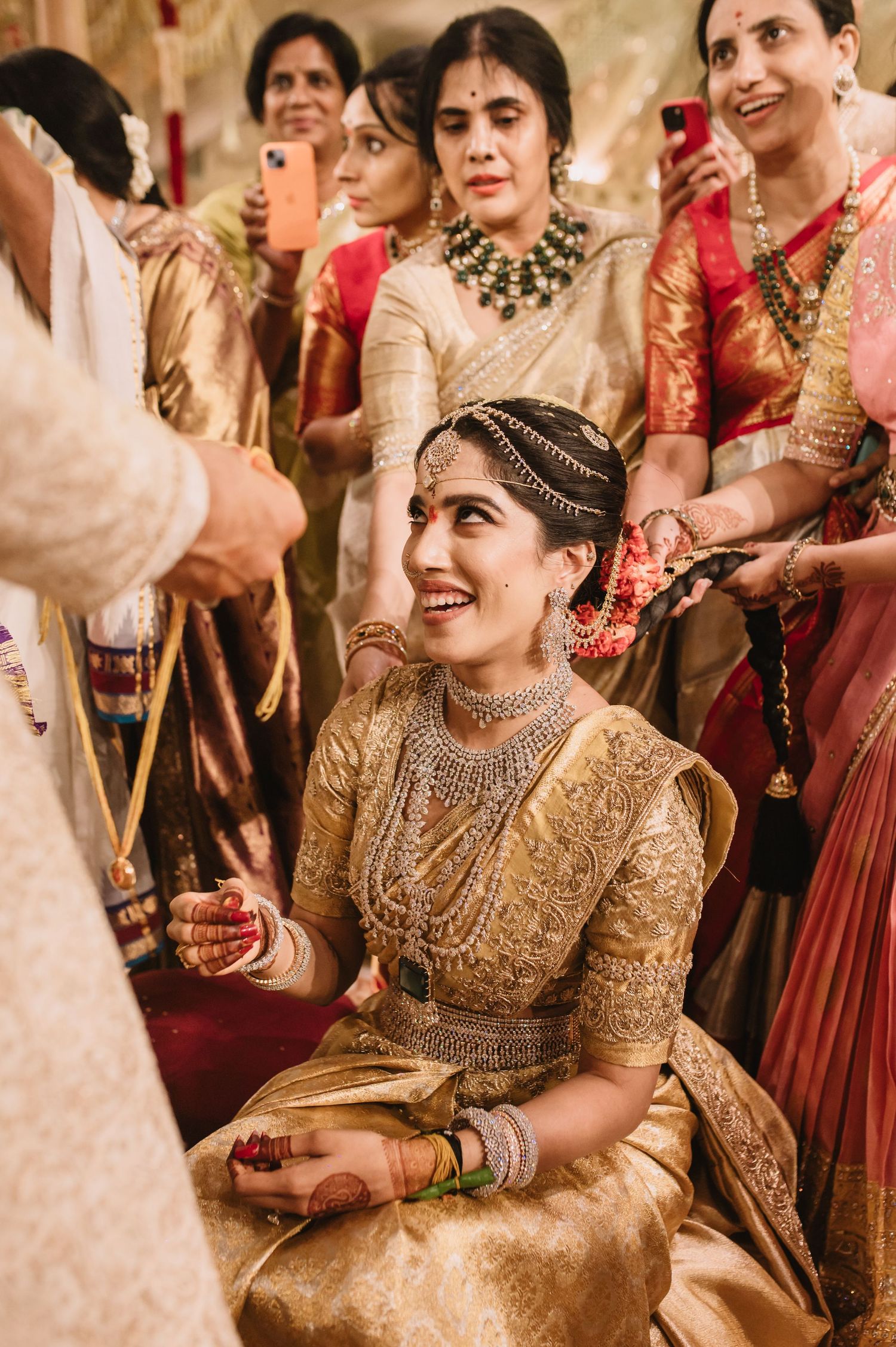 A smiling South Indian bride in gold silk saree with diamond jewelry sits during traditional wedding ceremony surrounded by guests.