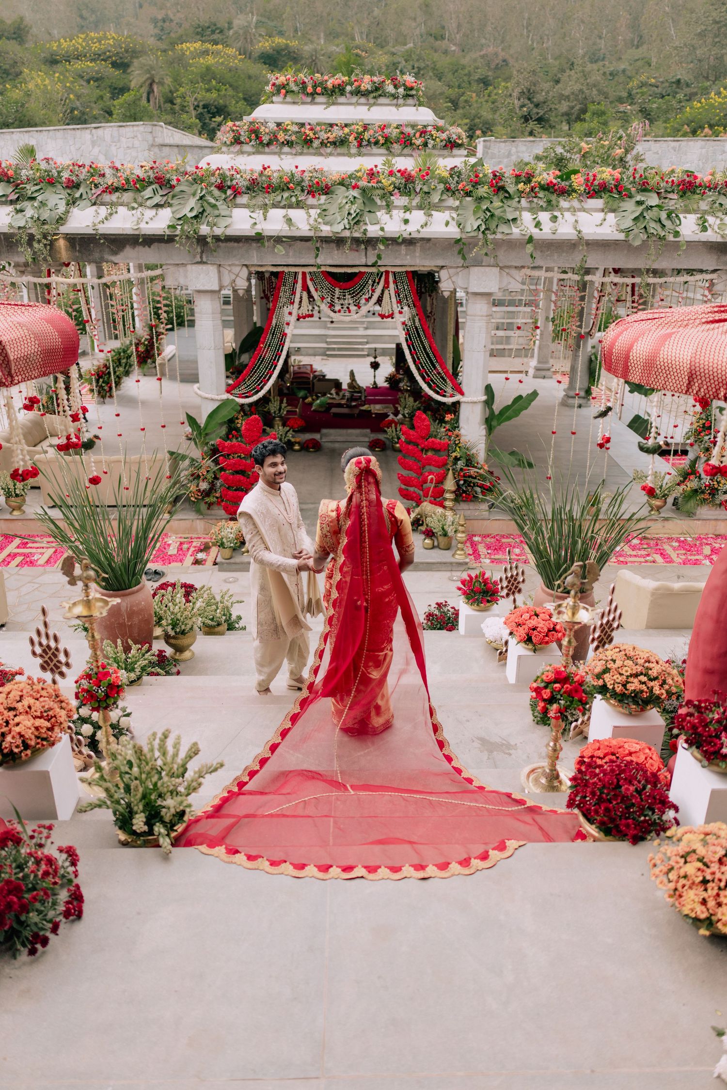 Bride in red lehenga with long veil walks with groom at ornate Indian wedding venue adorned with floral decor.