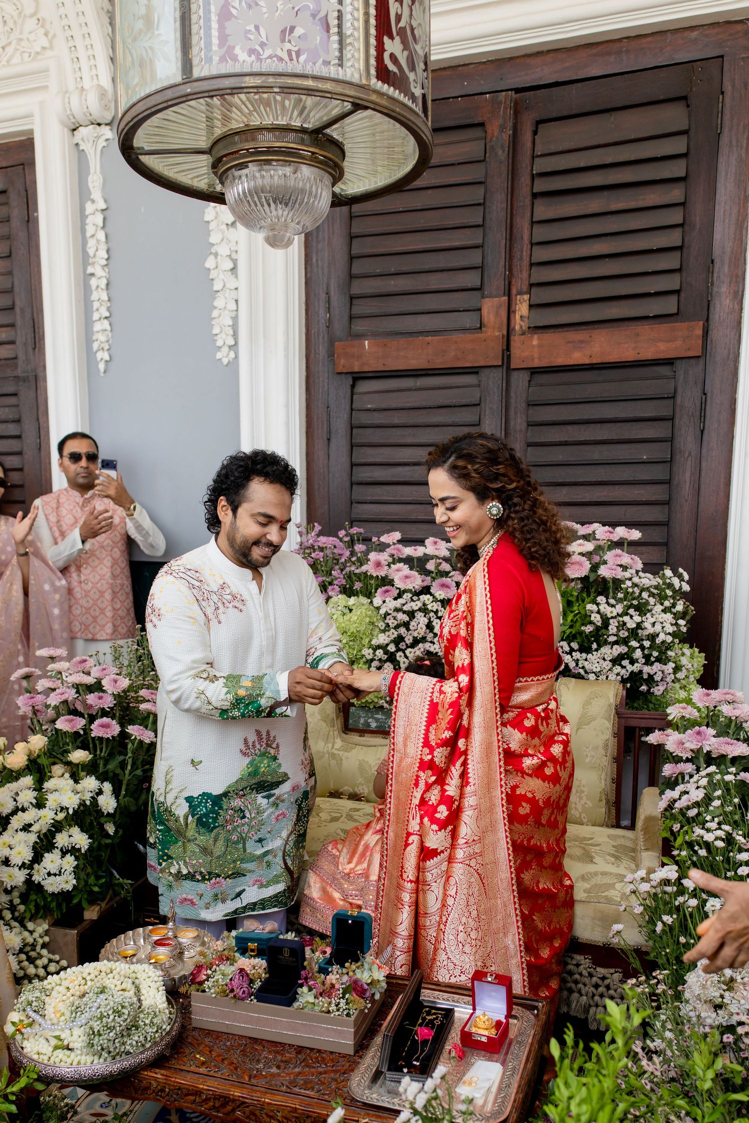 Indian couple exchanging rings during traditional wedding ceremony, bride in red saree, groom in white sherwani, floral backdrop.