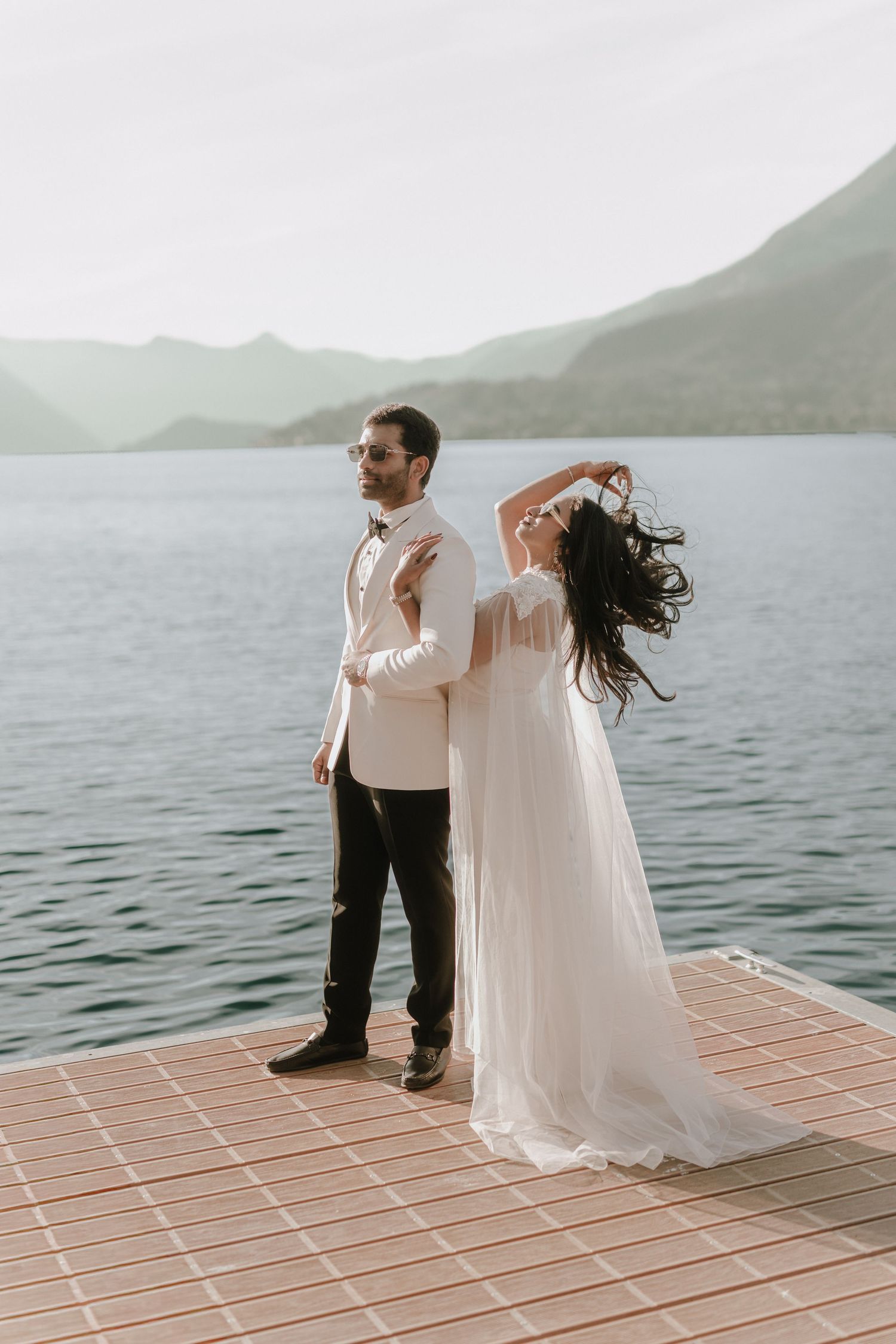 Bride in flowing white gown embraces groom on lakeside dock with misty mountains in background.