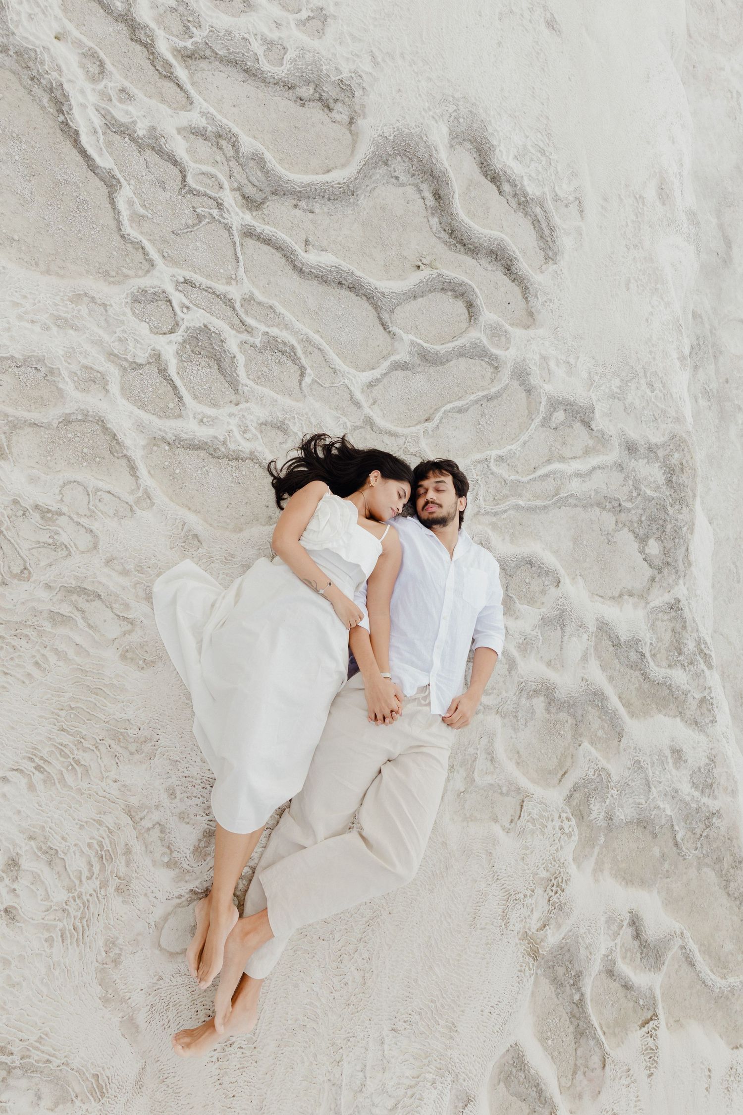 Aerial view of couple in white clothing lying together holding hands on white sandy terrain.