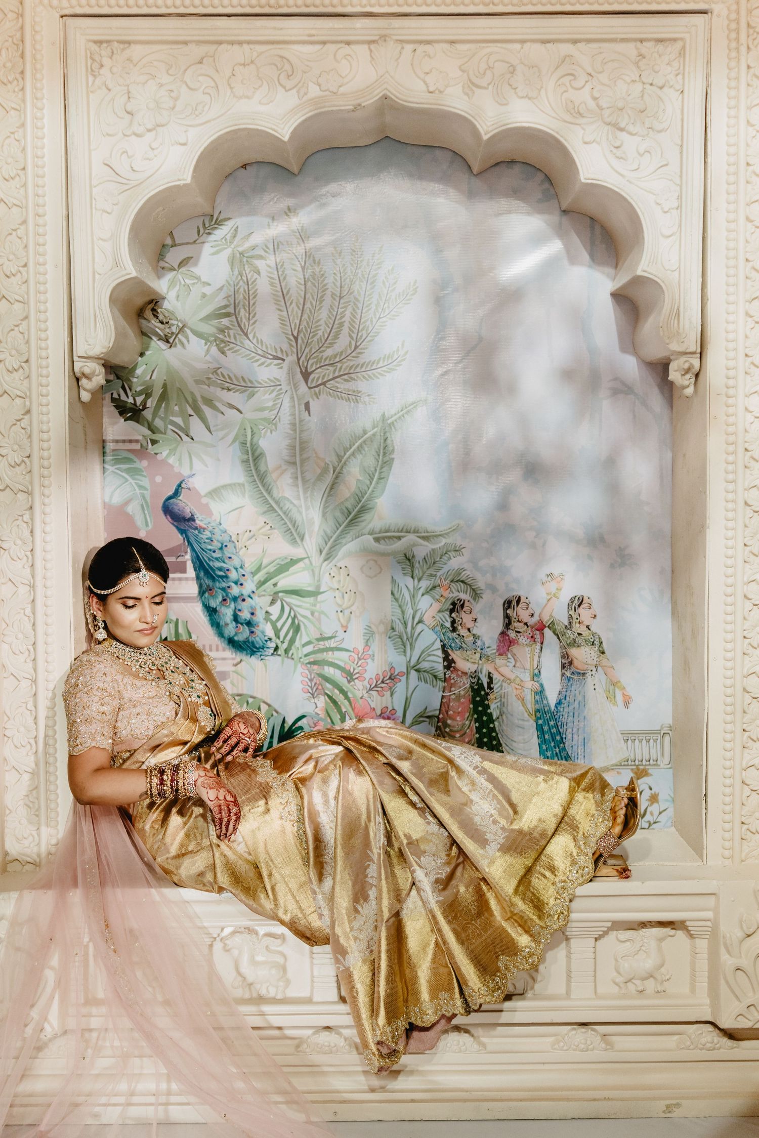 Radiant Indian bride in golden silk saree sits gracefully in ornate white arched alcove with floral mural.