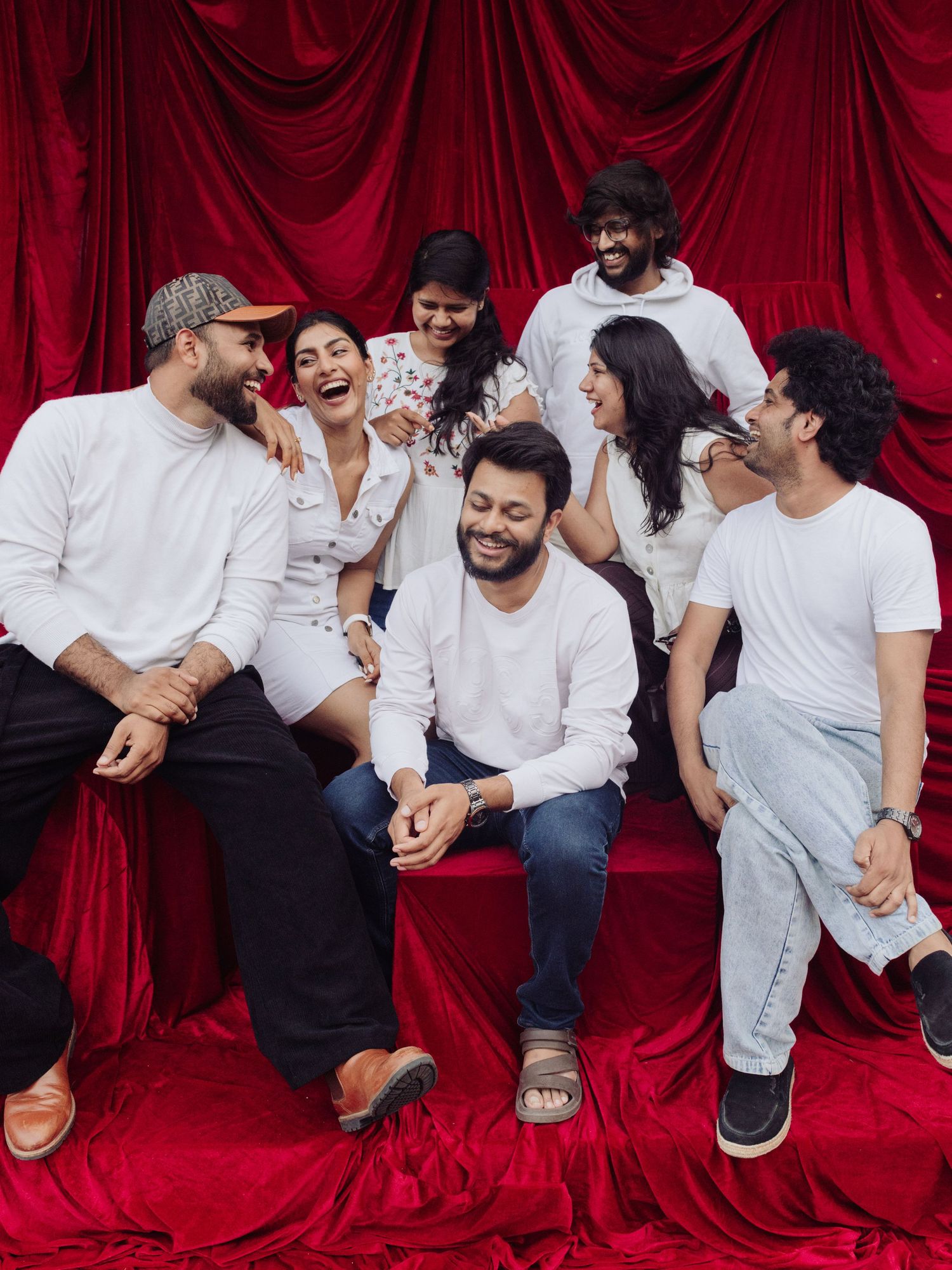 A group of seven friends laughing and posing together in white outfits against a rich red velvet backdrop.
