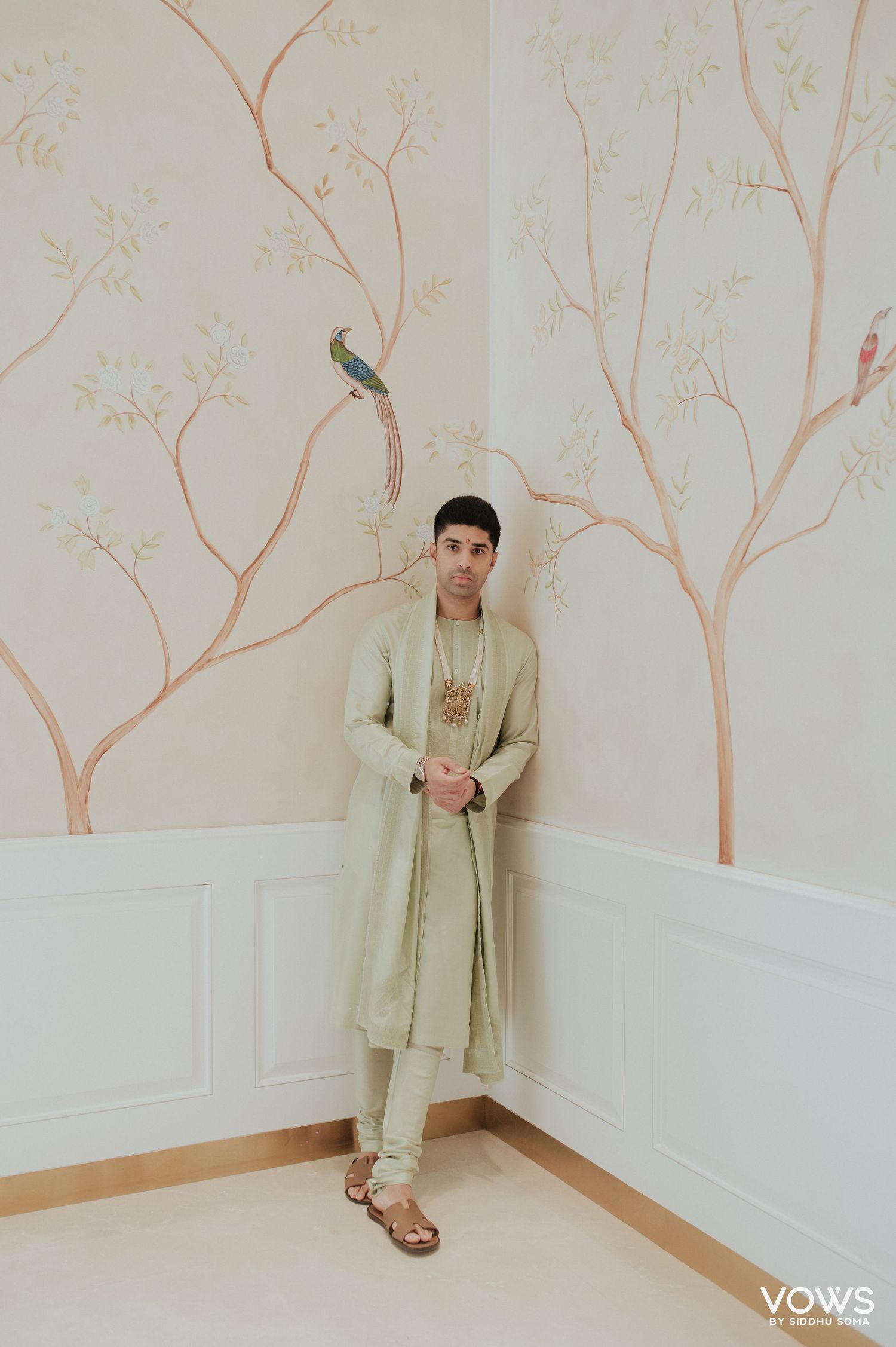 Groom in sage green sherwani poses against floral mural wall with painted birds and branches.