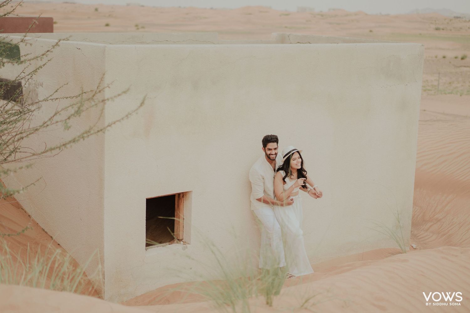 Couple posing together against a sandy desert wall in white outfits during a romantic pre-wedding photoshoot in Dubai.