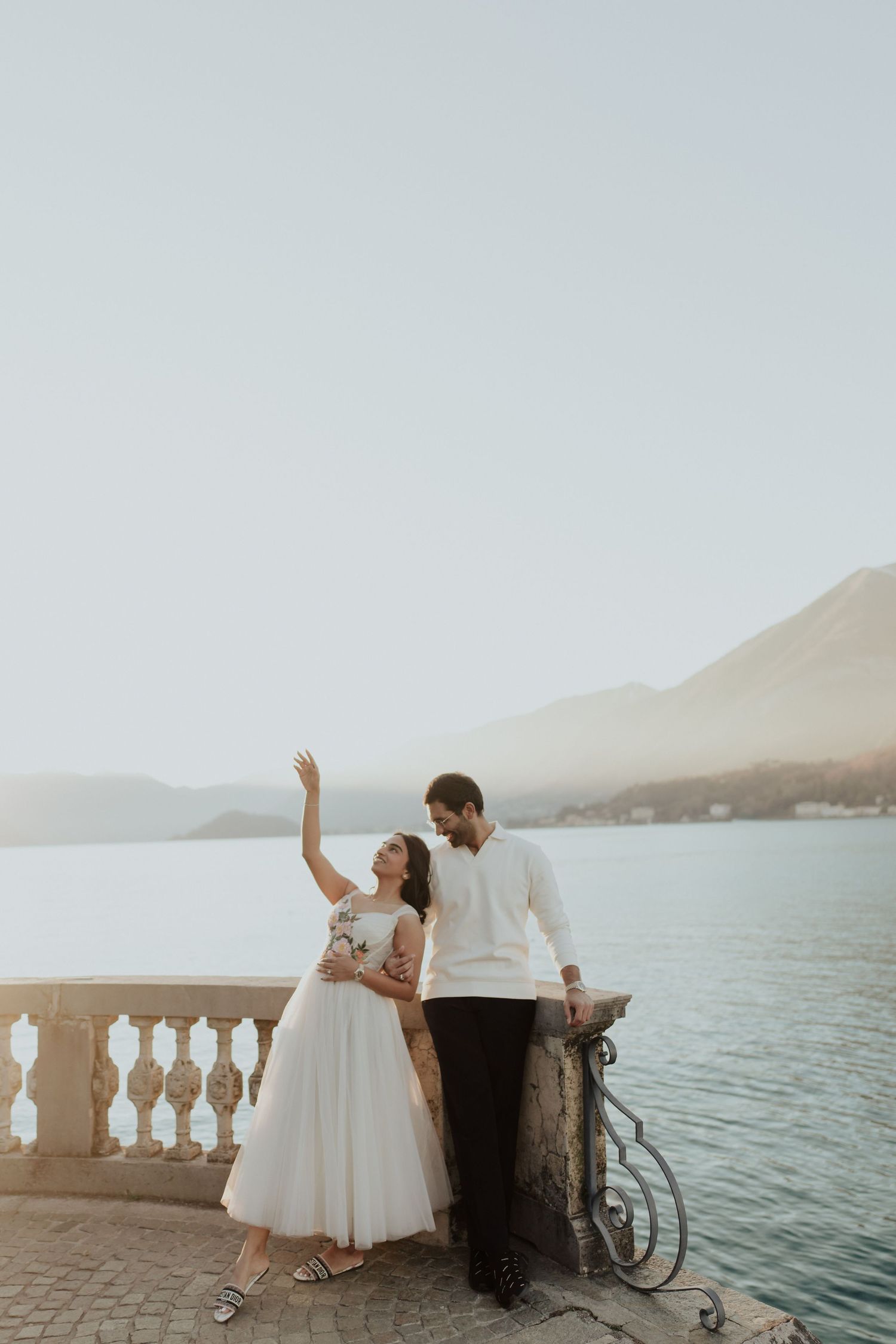 A bride and groom pose on a lakeside terrace with mountain views, she waves while he leans on a stone balustrade.