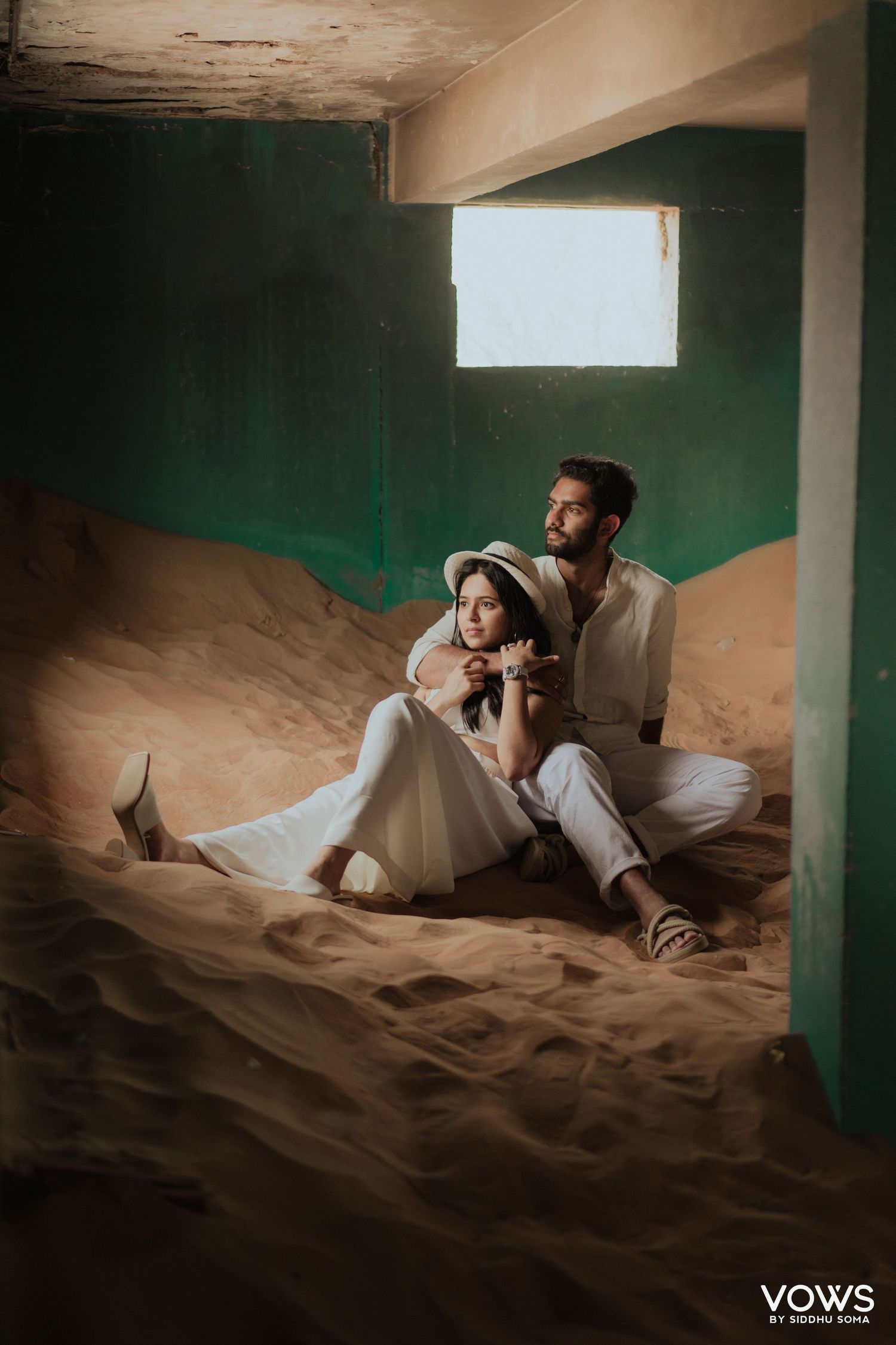 Couple in white outfits sitting on sand inside an abandoned building with green walls and a small window.