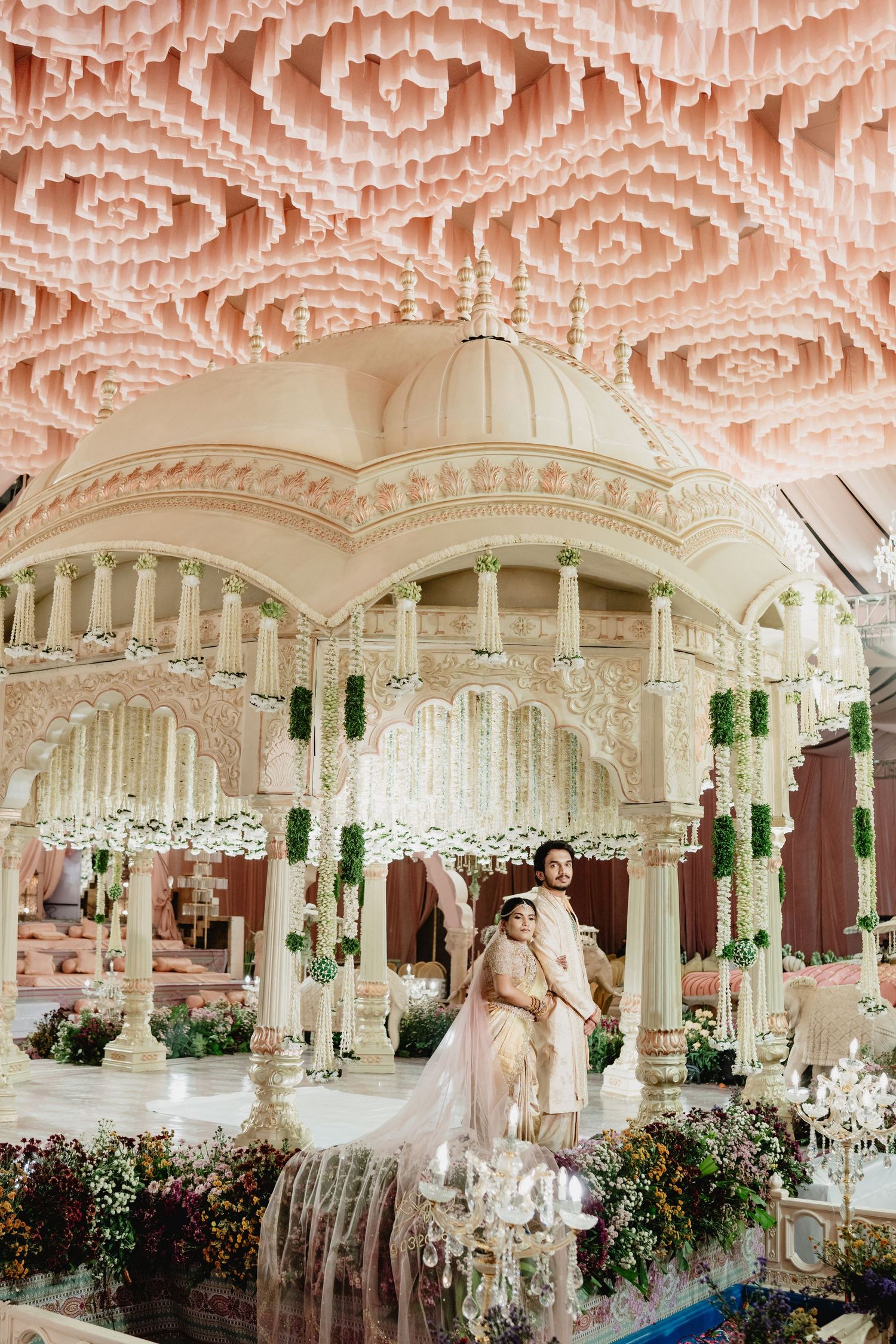 Bride and groom pose inside ornate white mandap with domed roof under pink rose ceiling installation at grand wedding.