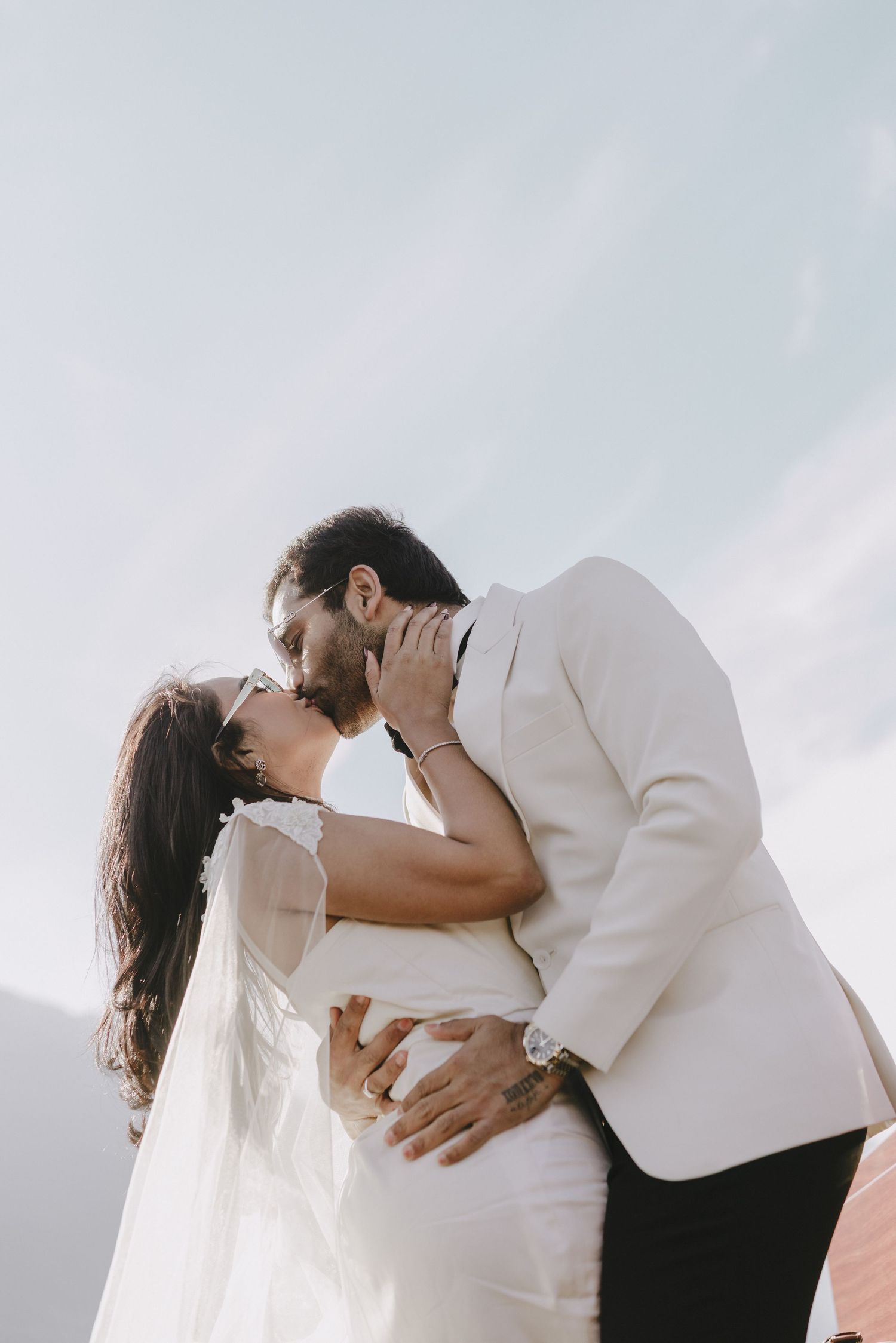 A couple shares a romantic kiss in wedding attire, bride in white gown with veil, groom in white suit, against a bright sky.