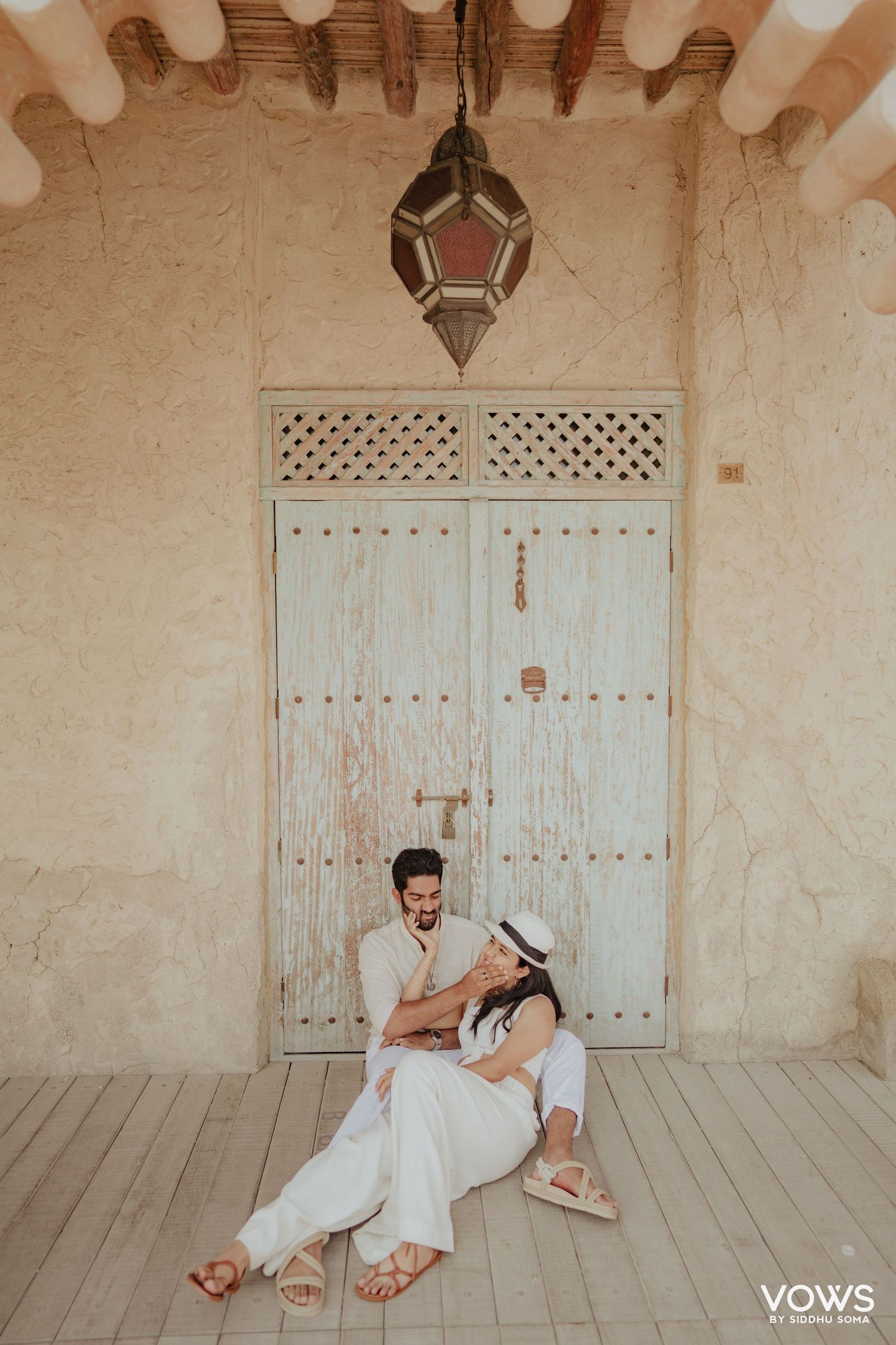Couple in white outfits sitting together by a rustic wooden door with Moroccan lantern overhead.