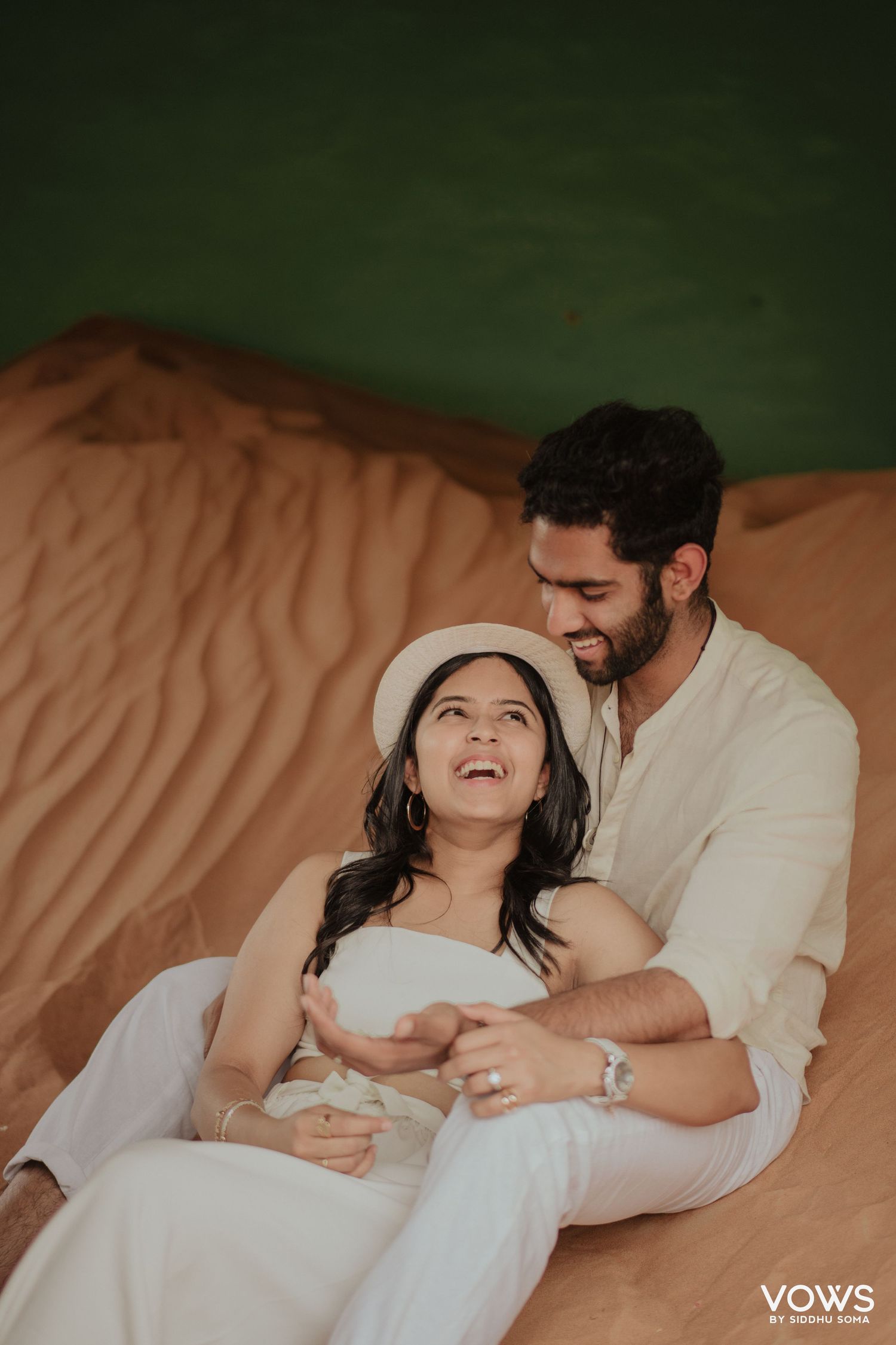 Couple sitting on sand dunes, woman in white dress and hat laughing joyfully while man smiles at her tenderly.