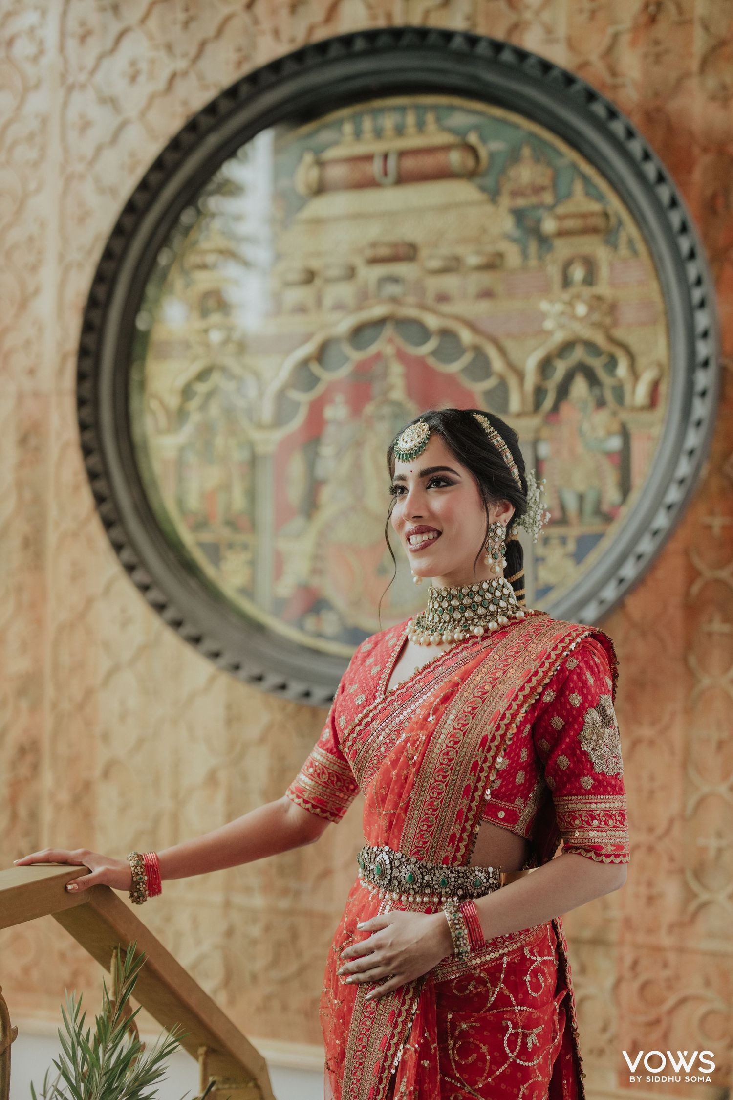 Bride in red embroidered saree with gold jewelry poses elegantly before ornate circular artwork backdrop.
