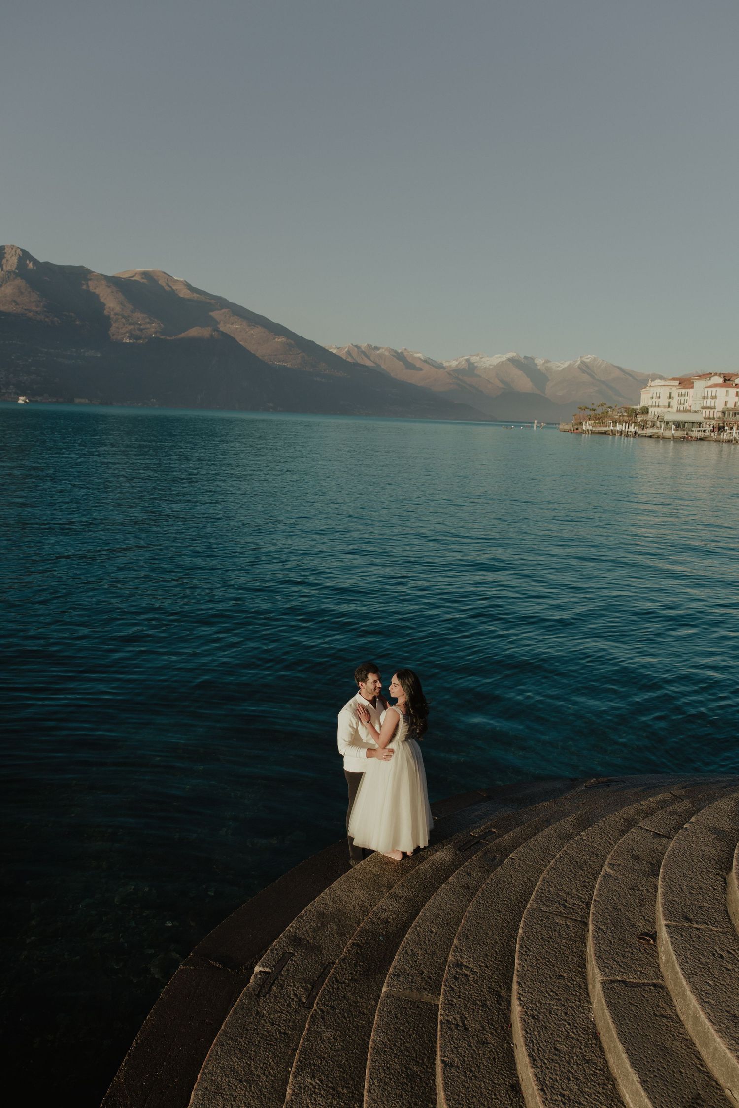 Bride and groom embracing on stone steps by a turquoise lake with mountain backdrop on their wedding day.