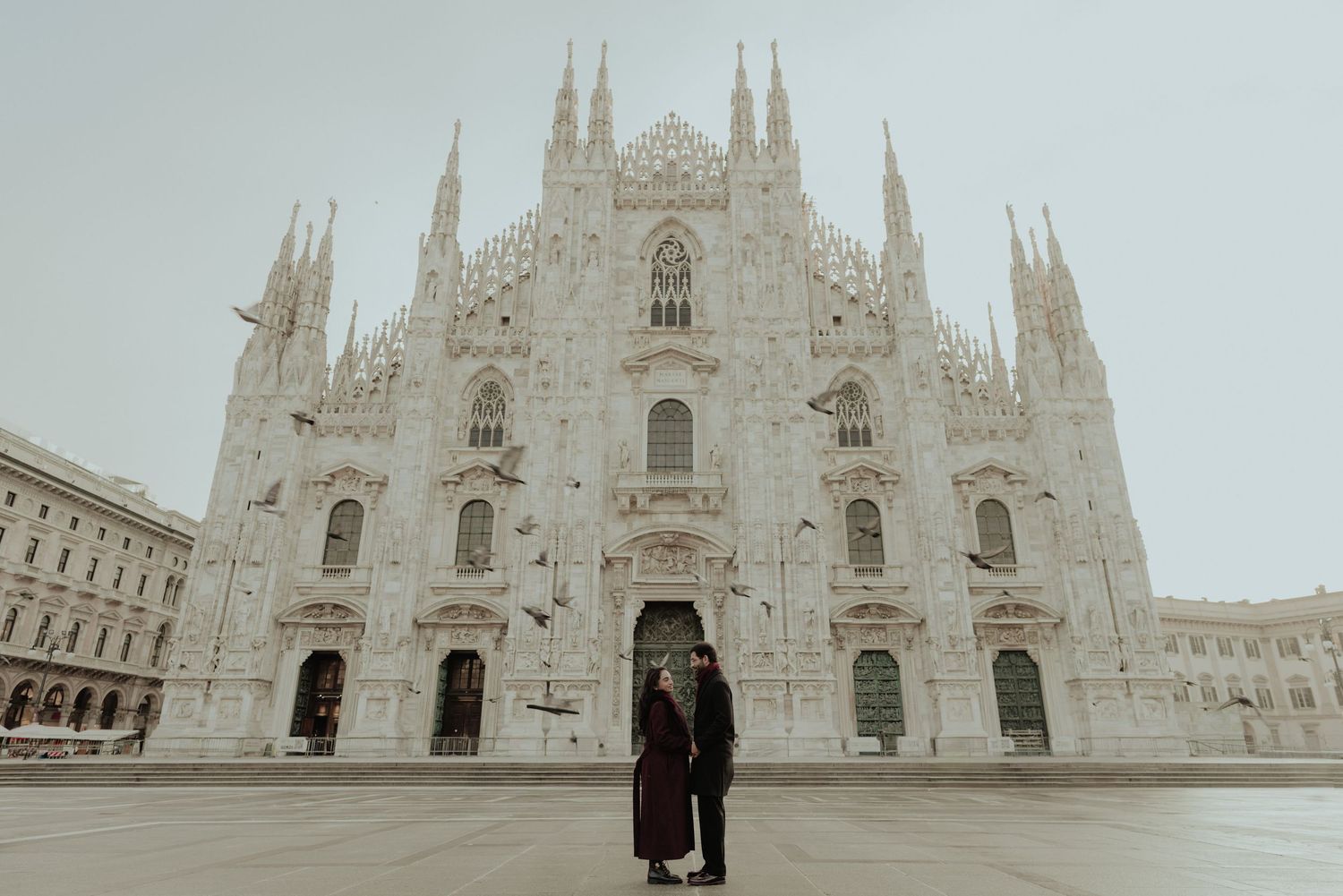 Couple standing hand in hand before the grand Gothic facade of Milan's Duomo cathedral.