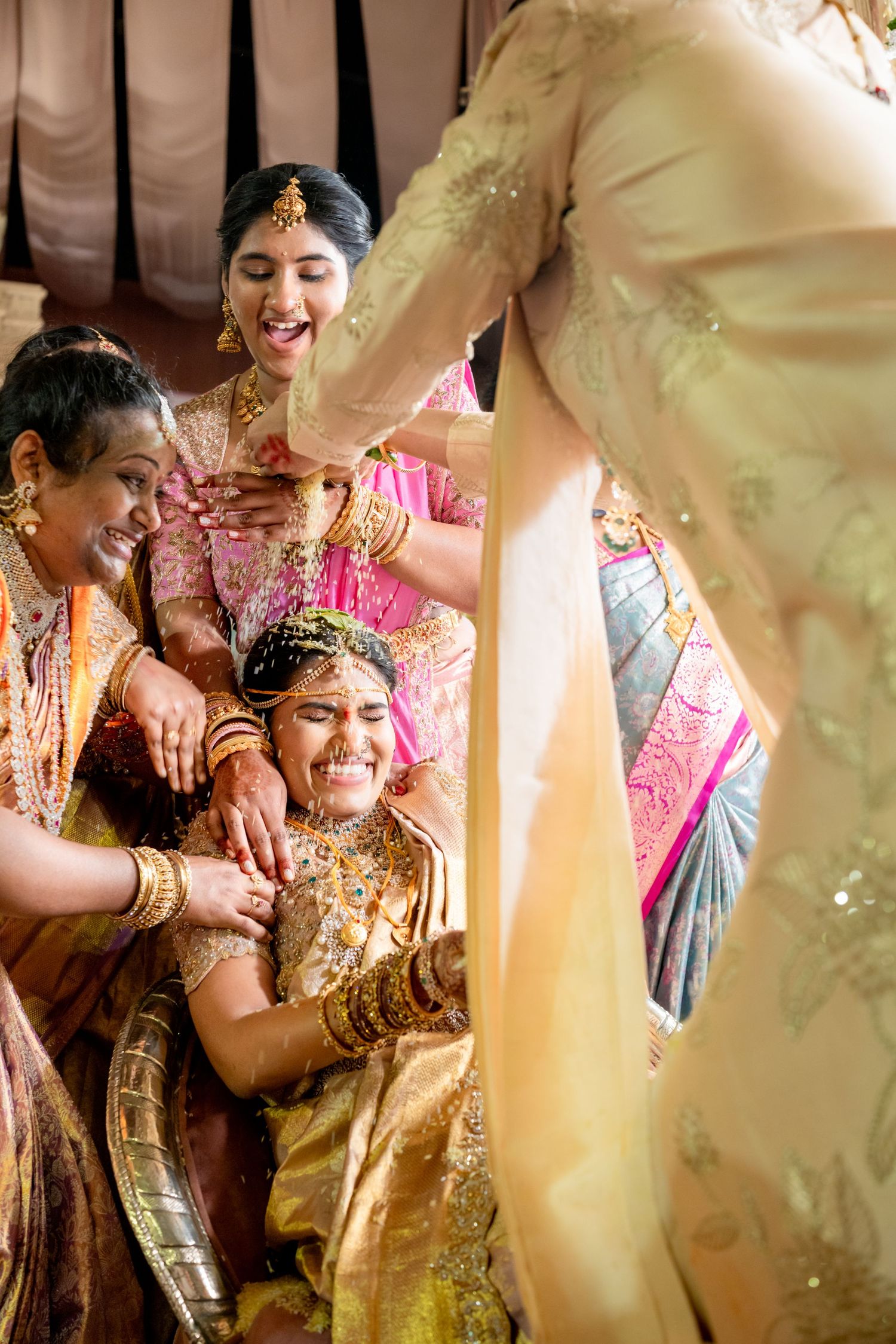 Female family members joyfully assist bride in golden silk saree during traditional South Indian wedding ritual.