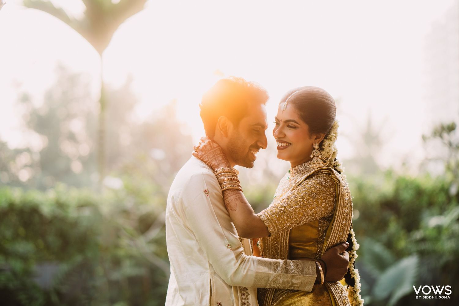 Indian couple in traditional attire sharing a joyful embrace outdoors with golden sunlight glowing behind them.