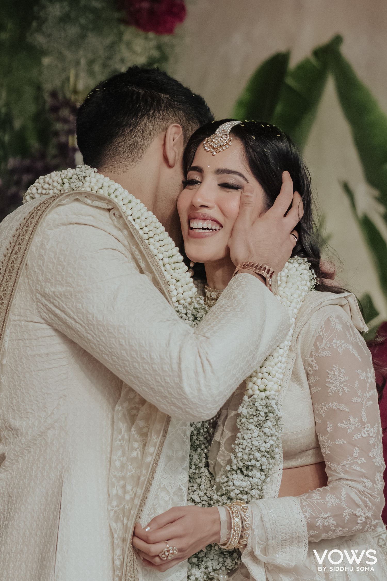 Couple in white traditional Indian wedding attire sharing a joyful moment, bride smiling as groom kisses her cheek.